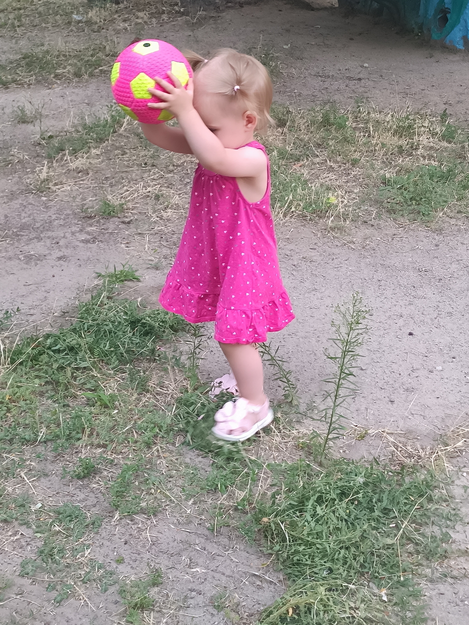 A young girl, likely a toddler, stands on a patch of dirt and grass, holding a pink and neon yellow soccer ball to her chest. She is wearing a bright pink dress adorned with small white hearts and pink sandals. Her blonde hair is pulled into two pigtails, each tied with a white elastic band. The girl appears to be looking down at the ball, her face mostly obscured.
The setting is outdoors, with dry dirt and patches of green grass and weeds. The lighting suggests it is daytime, though the exact time is unclear. The background is out of focus, showing more of the ground and a hint of something colorful and possibly painted in the upper right corner. There is no visible text in the image. The overall scene depicts a moment of play or perhaps curiosity with the soccer ball.