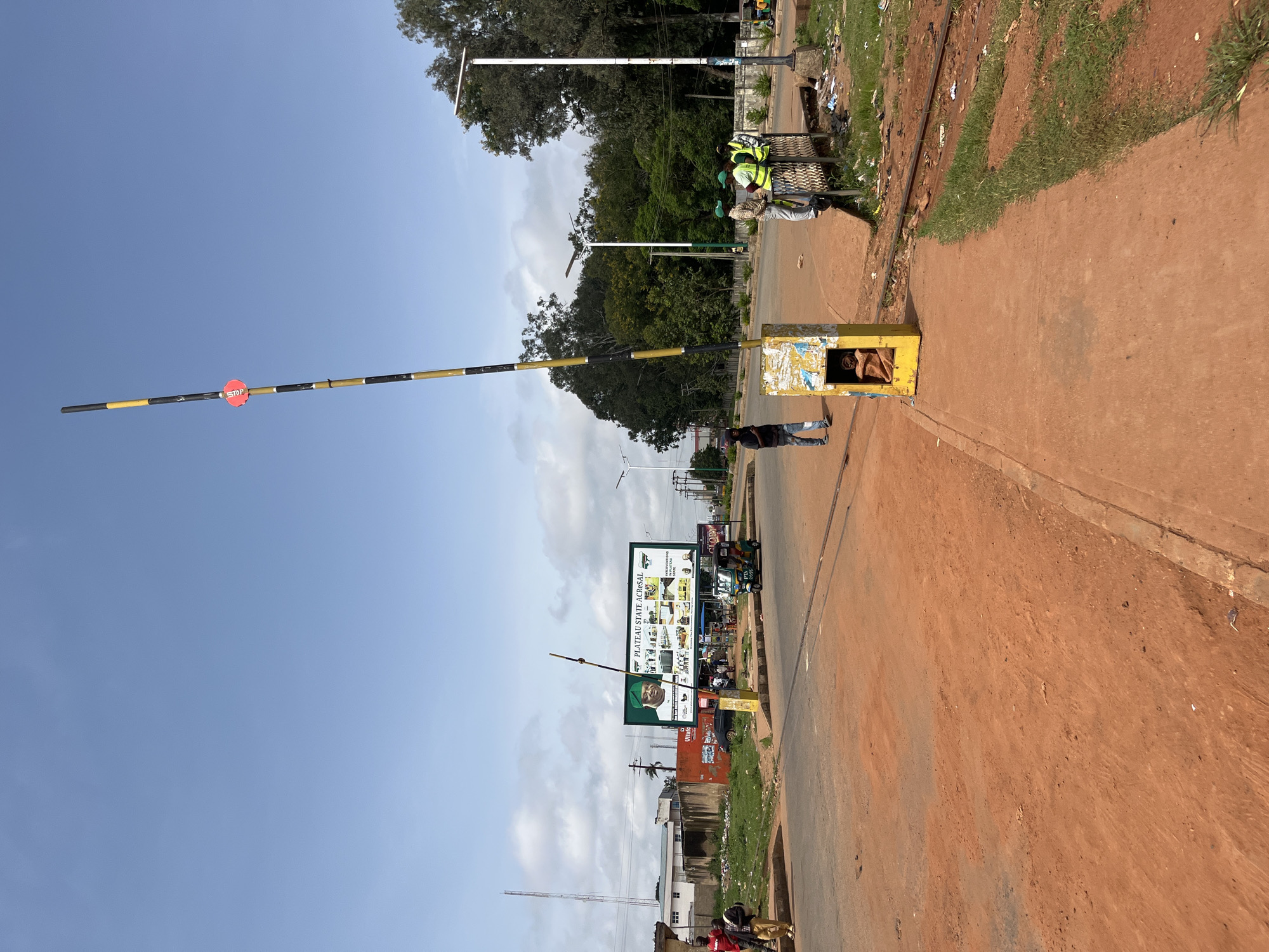 A barrier arm, striped black and yellow with a red stop sign, is lowered across a dirt road in Bukuru, Nigeria. The barrier is mounted on a yellow, concrete base. In the background, a billboard displays images and text related to Plateau State. Several people, some wearing high-visibility vests, stand near the barrier, conversing. A trishaw is parked to the right of the barrier. The ground is covered in reddish-brown dirt, and there is evidence of trash and debris in the immediate vicinity. The sky is clear blue with some scattered clouds. The scene appears to be a road crossing or checkpoint.