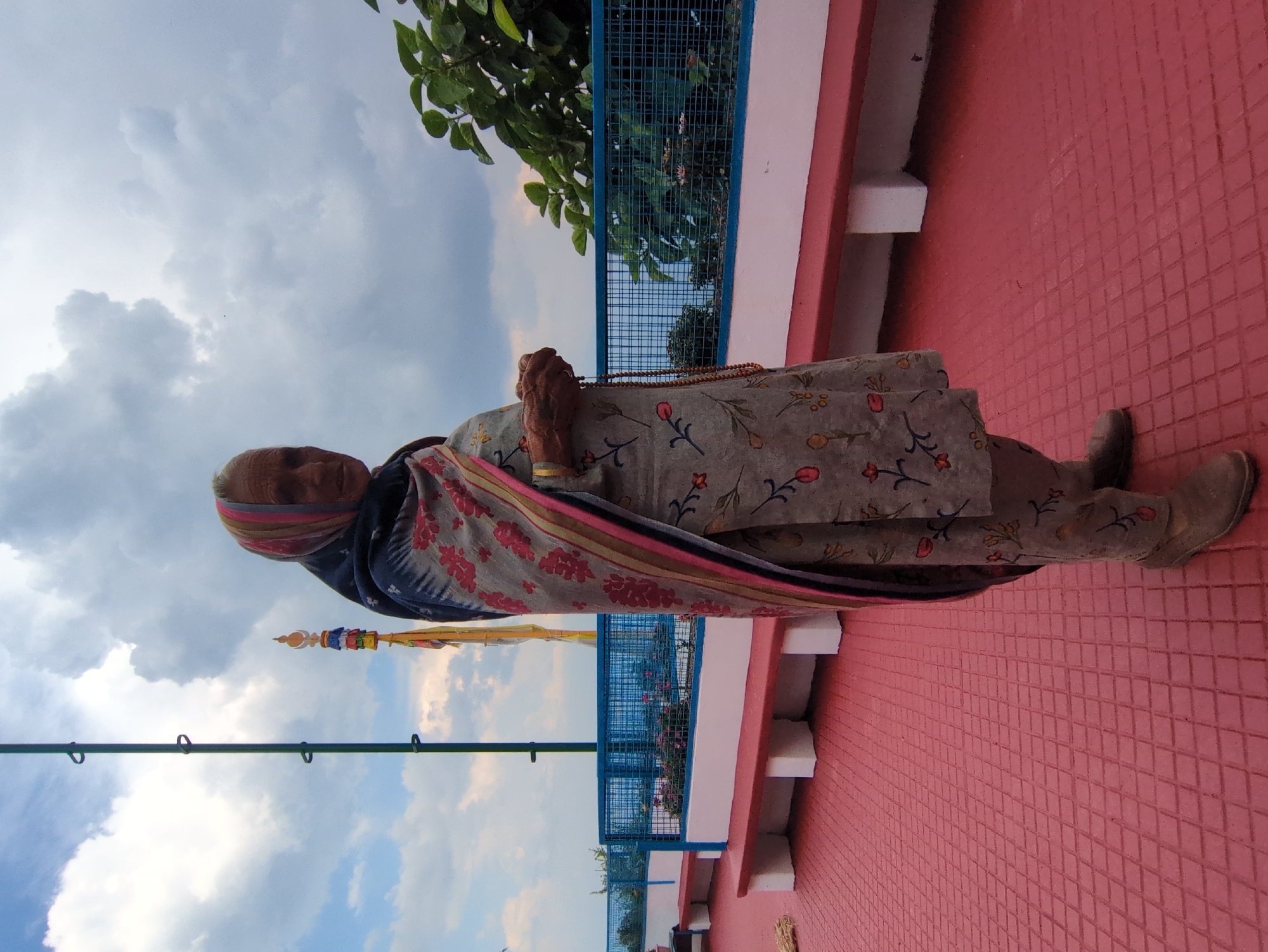 In Doh, India, an elderly woman stands on a red tiled patio, facing right and looking towards the camera. She is wearing a floral patterned grey kurta and pants, with a colorful striped shawl draped over her shoulders and head. Her hands are clasped together, holding a string of prayer beads. Behind her, a blue metal railing encloses a garden area with white benches. The sky above is cloudy with patches of blue. A tall green pole with several rings stands to the left of the woman, and a colorful, tiered religious structure is visible in the distance. Lush green foliage is present on the right side of the frame.