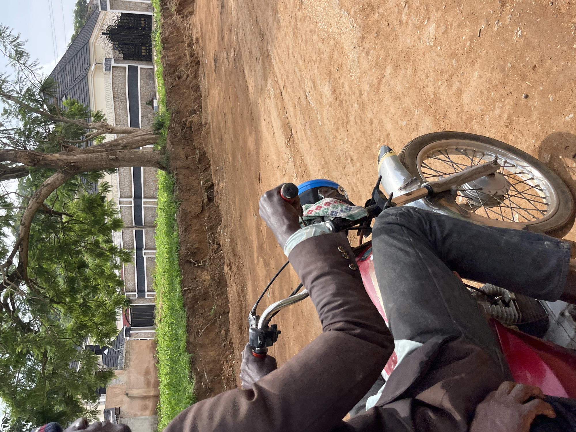 In Bukuru, Nigeria, a person in a brown suit and dark pants rides a red motorcycle on a dirt road. The person's hands are on the handlebars, with one hand gripping a red throttle. The motorcycle has a silver fender and a visible engine. In the background, a stone and stucco wall with a black metal gate encloses a modern building with a dark roof, surrounded by green foliage and trees. The road is unpaved, with visible tire tracks and scattered stones. The scene appears to be outdoors during daylight.