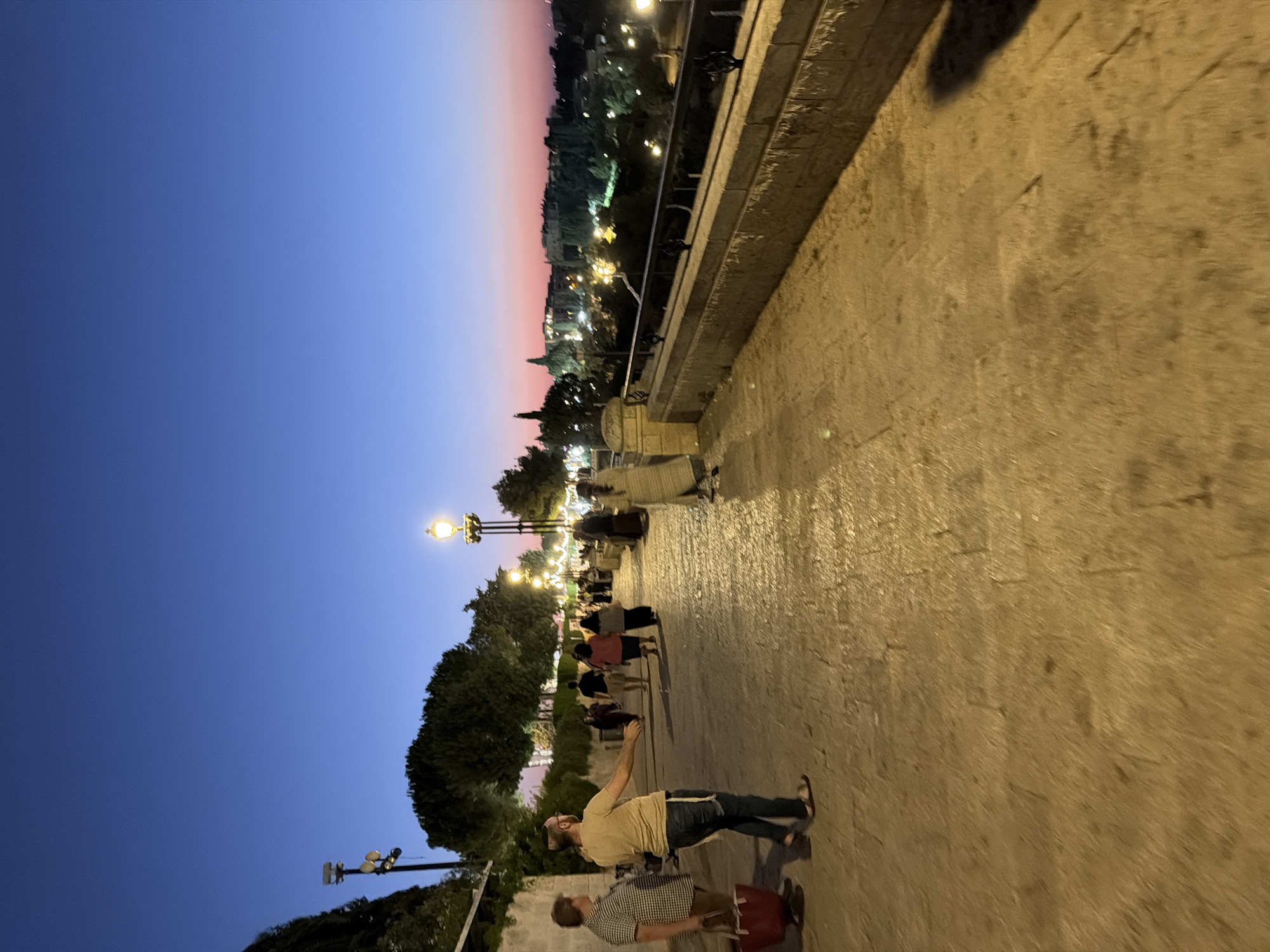 The image captures a twilight scene along a paved pathway in Jerusalem, Israel, likely overlooking the Old City. The sky transitions from a deep blue overhead to hues of pink and orange near the horizon, indicating sunset or dusk. Several people are walking along the pathway. In the foreground, two men are interacting; one, wearing a light-colored shirt and jeans, gestures with his arm, while the other, in a plaid shirt, holds a red bag. Further along the pathway, other individuals, some in traditional or modest attire, are walking or standing. Lampposts illuminate the scene, casting a warm glow on the stone paving. The pathway is bordered by a stone wall with decorative metal railings, beyond which a cityscape with illuminated buildings and trees is visible. Lush trees line the left side of the pathway, and a stone wall marks the edge of the elevated area. The overall atmosphere is serene and suggests a moment of shared experience in a historic setting.