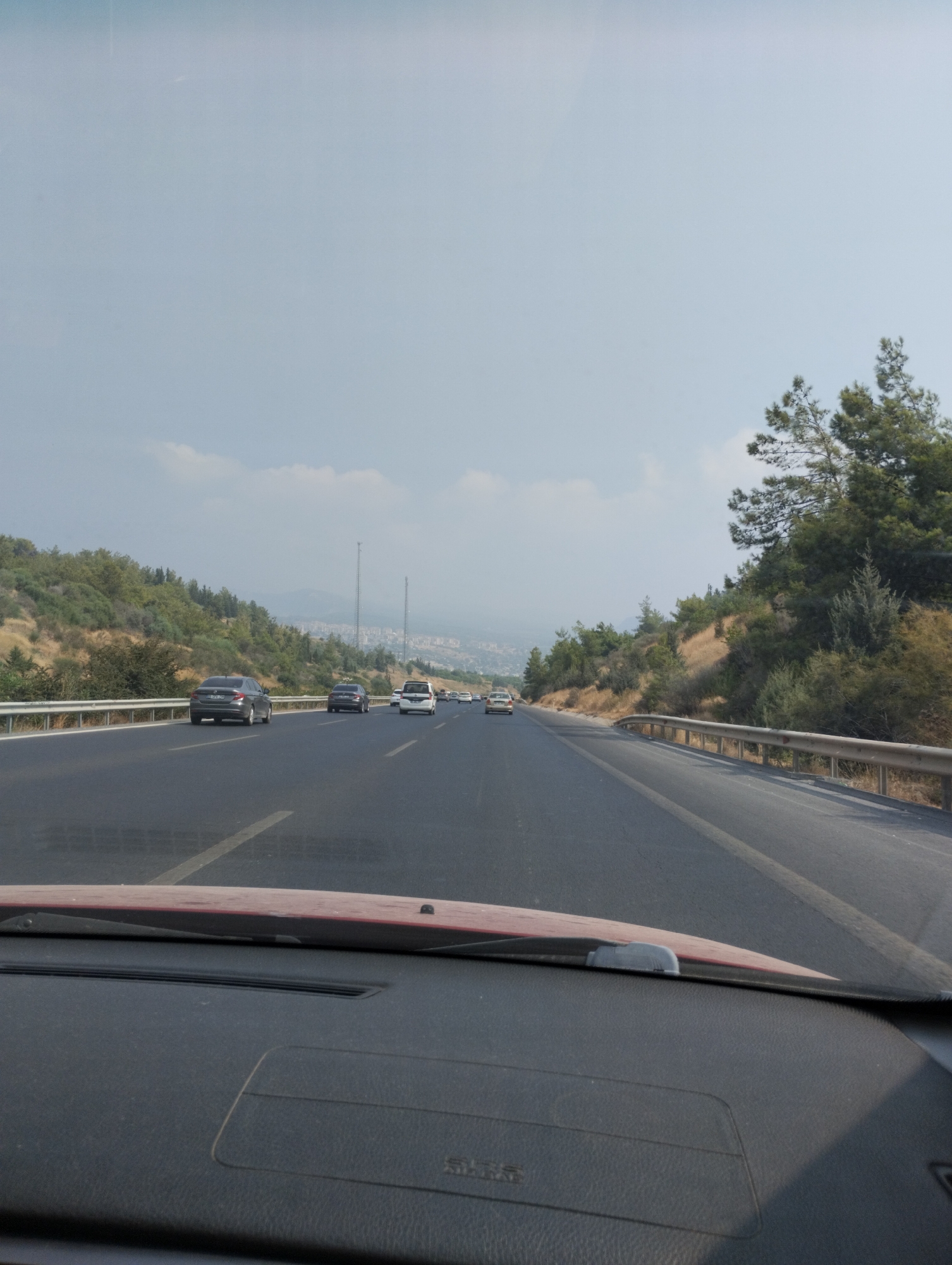 The image captures a view from inside a vehicle driving on a highway. The highway has multiple lanes and is flanked by dry, grassy hills dotted with trees and shrubs. In the distance, a city with buildings is visible under a hazy sky with scattered clouds. Several cars are driving on the highway in the same direction as the viewer. The foreground shows the dashboard and windshield of the vehicle. The scene is consistent with a drive on the coastal highway near Mezitli, Turkey, likely during the daytime.