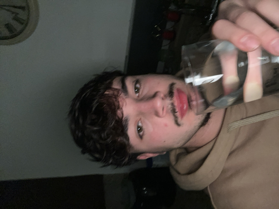 A person with dark curly hair and a mustache is holding a glass of water to their mouth. They wear a tan hoodie. Behind them, a wall is visible with a clock hanging on it. Other items are blurred in the background, including what appears to be a red object. The image has a low-light setting, creating a dim ambiance.