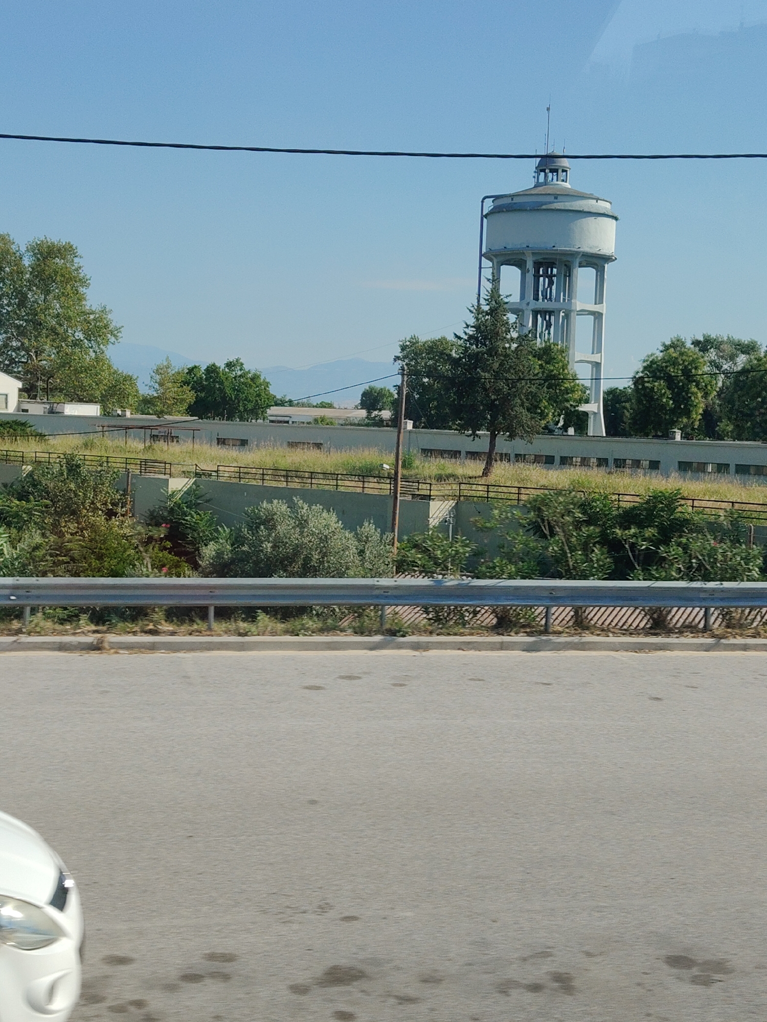 This image depicts a water tower in Larissa, Greece, on a clear day with a blue sky. The water tower is a tall, cylindrical structure with a domed roof, supported by a multi-legged concrete base. Surrounding the water tower are various trees and vegetation, and in the foreground, a road and a guardrail are visible. The scene appears to be a street-level view, possibly from a vehicle, as suggested by the angle and the presence of a car headlight in the lower-left corner of the frame. The overall atmosphere is serene and rural, with the water tower standing as a prominent landmark against the backdrop of the natural landscape and the distant mountains.