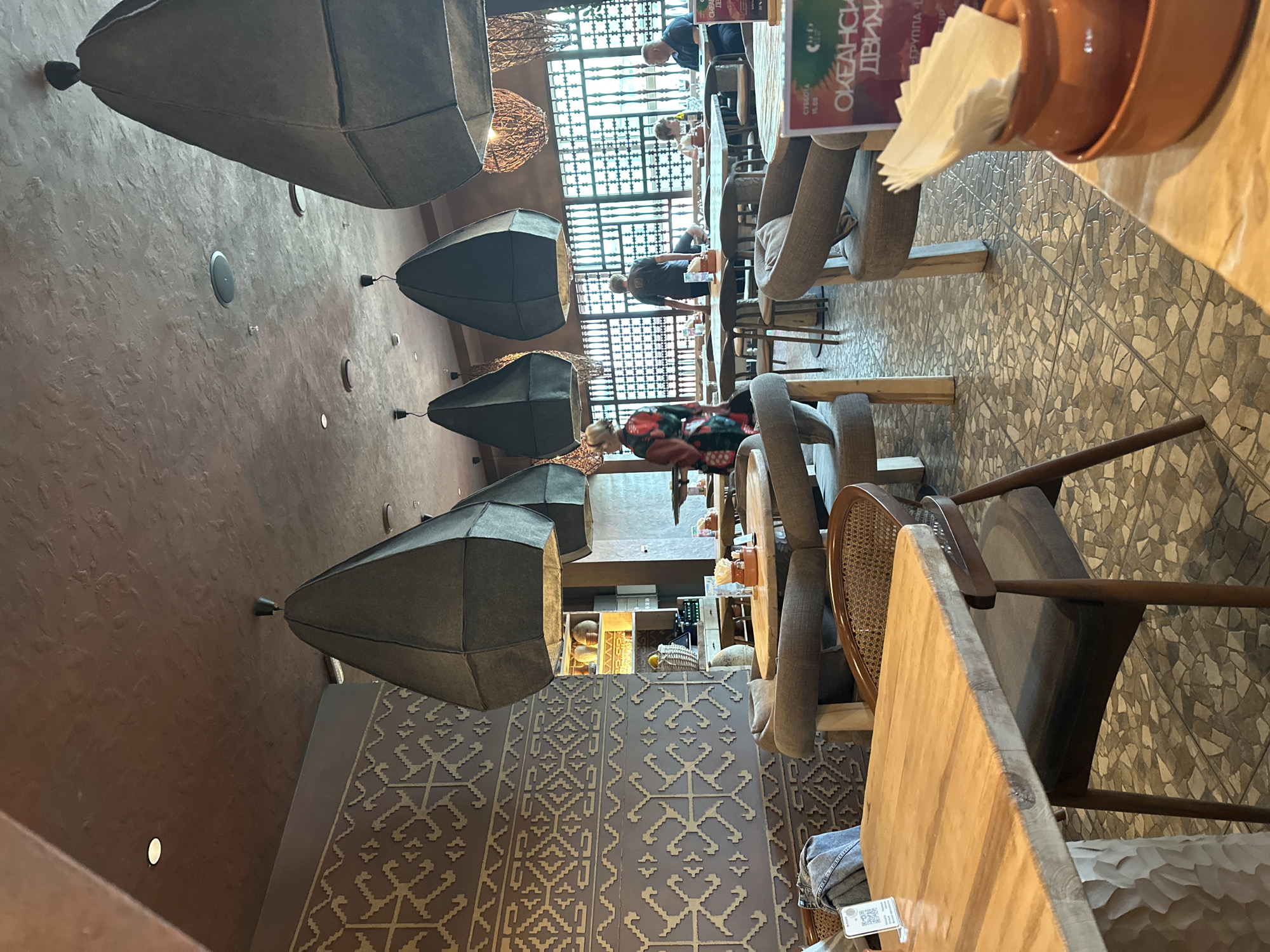 The image shows the interior of a restaurant. Several large, dark grey, geometric lamps hang from the brown, textured ceiling. The walls are adorned with a decorative pattern of light beige geometric shapes against a darker background. Tables and chairs are arranged throughout the space. In the foreground, a wooden table and chairs are visible. Further back, several people are seated at tables or walking. One person, dressed in a colorful robe, walks from left to right. Another person in a dark shirt stands near a window. The floor is tiled with a mosaic of grey and beige irregular shapes. In the mid-ground, a decorative screen or window structure is visible behind the tables. The overall atmosphere appears to be dimly lit and cozy. The image is of a restaurant located in Petropavlovsk-Kamchatsky, Russia.
