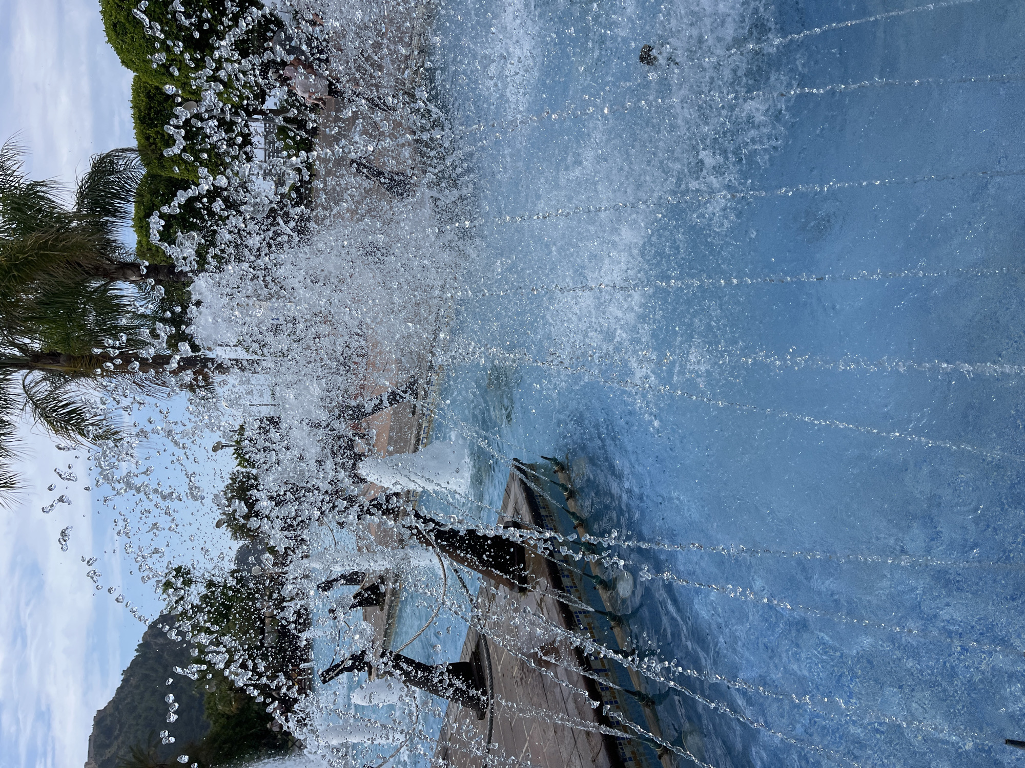 The image captures a view through a fountain in Alanya, Turkey. The water droplets from the fountain are prominent in the foreground, obscuring and partially distorting the background.
Behind the fountain, a partially visible mountain covered in green vegetation is present on the left side of the frame. A few palm trees and dense greenery are also visible. The water in the fountain is a pale blue color. A structure, possibly the fountain's base or a nearby building, is partially visible behind the water streams.