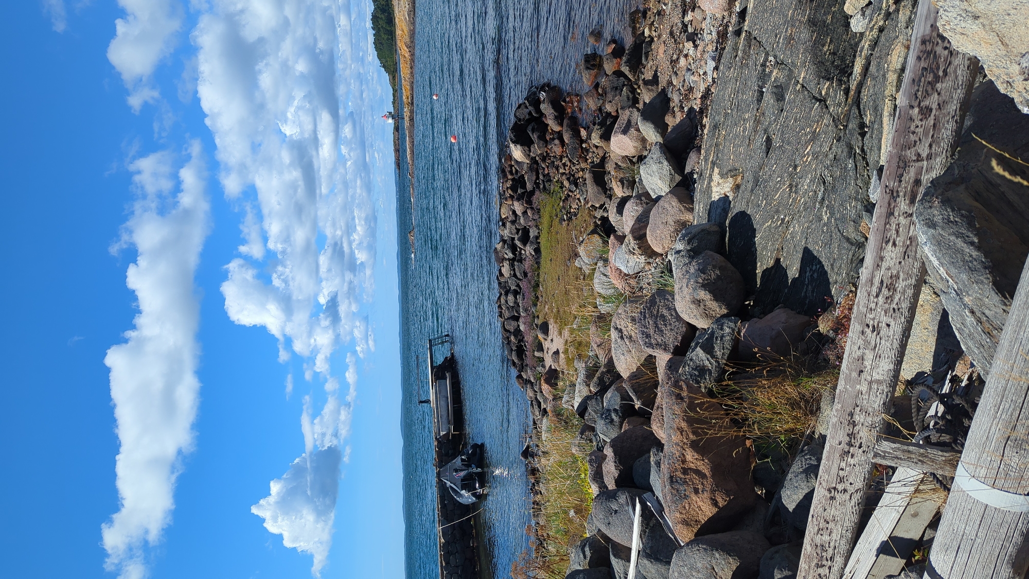 The image depicts a rocky shoreline in Larkollen, Norway, under a bright blue sky with scattered white clouds. In the foreground, weathered logs lie on the stones. The middle ground features a rocky embankment leading to the water. A stone jetty extends into the calm, dark blue sea, where a small boat is moored. Adjacent to the jetty is a wooden diving platform. Further out in the sea, two orange buoys are visible, and a red and white lighthouse stands on a rocky outcrop in the distance, situated on a tree-covered headland. The overall scene is one of a serene coastal environment.