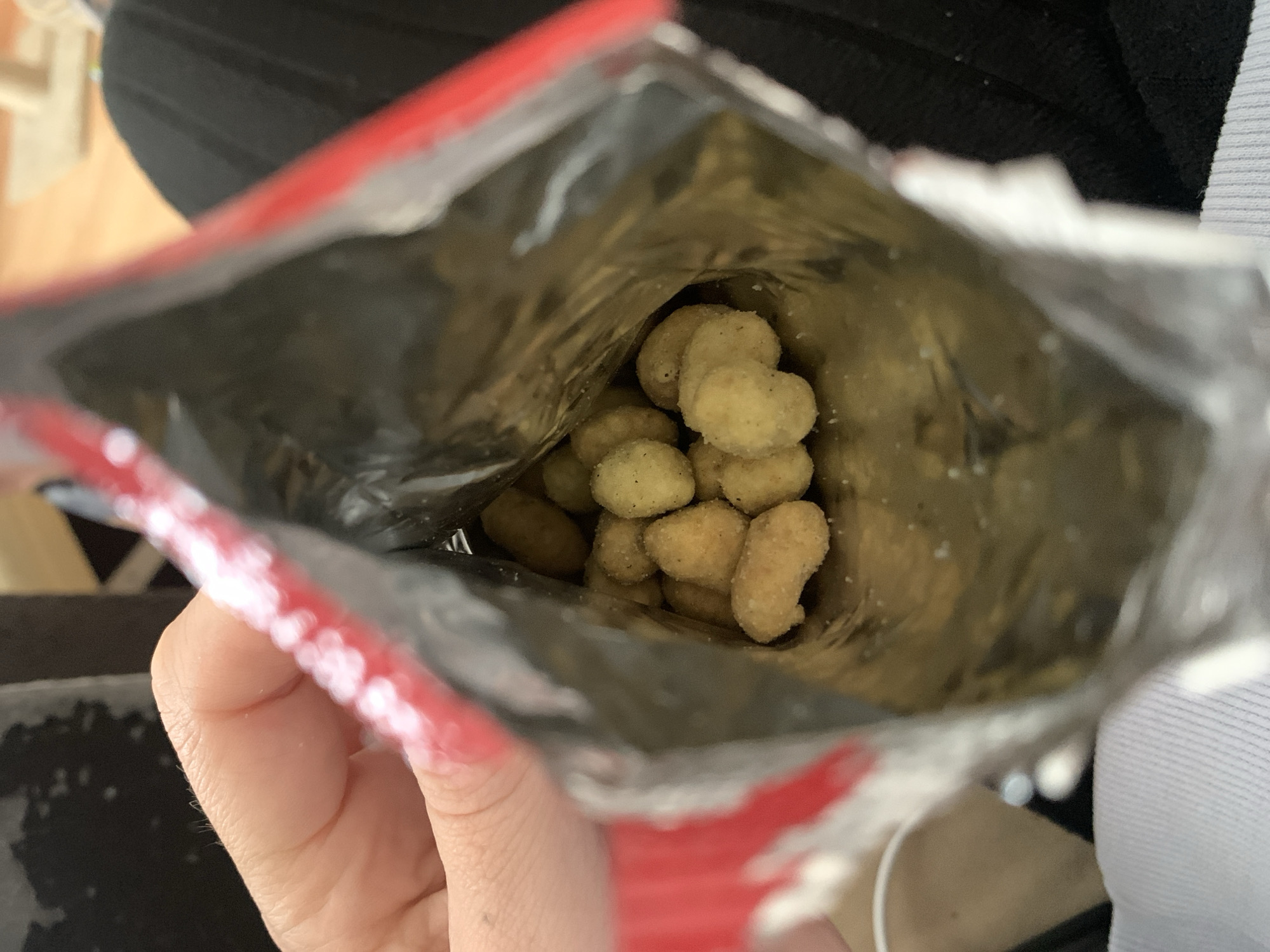 A close-up, top-down view captures the inside of an open snack bag held by a person's hand, likely in Stavropol, Russia. The bag contains numerous small, irregularly shaped snacks, predominantly light brown or beige, and coated with a visible seasoning or powder. Some pieces have a distinct heart-like or nugget shape. The interior of the snack bag is shiny and silver, suggesting a foil lining, while the visible edge of the bag's opening is red. A person's fingers are visible, gripping the base of the bag, implying the bag is being held upright and tilted to access the contents. The background is out of focus, showing indistinct shapes and colors, including a light-toned surface (possibly wood) on the left and dark fabric on the right, indicating an indoor, casual setting. The activity depicted is snacking. No discernible text is visible on the snack packaging or within the frame. The time of day and specific weather conditions are not ascertainable from the indoor view.