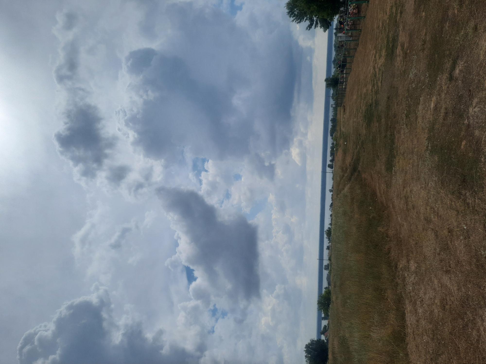 The image depicts a wide landscape under a cloudy sky. In the foreground, there is a dry, grassy field with a dirt path winding through it. The middle ground features a body of water, likely a river or lake, with a distant treeline and some scattered buildings along the shore. The sky is dominated by large, dramatic clouds, with patches of blue sky visible. There is a fence running along the right side of the middle ground, with some structures or objects behind it. No people or animals are visible in the image. The overall impression is one of a rural or semi-rural setting, possibly a cemetery, given the fence and potential markers visible behind it. The location is identified as Крестьянка, Russia.