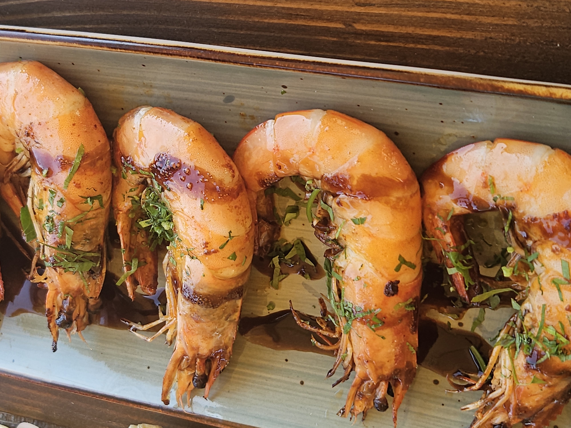 Four large, grilled prawns are arranged vertically on a rectangular, muted green ceramic plate. The prawns are cooked to a vibrant orange hue with visible grill marks and a glossy, dark sauce drizzled over them. They are garnished with finely chopped green herbs, likely parsley or chives. The plate rests on a dark wooden surface, suggesting a table setting. The lighting is bright, creating highlights on the prawns and their sauce. There are no people visible in the image. The scene appears to be a close-up food photograph taken in Jošice, Montenegro.