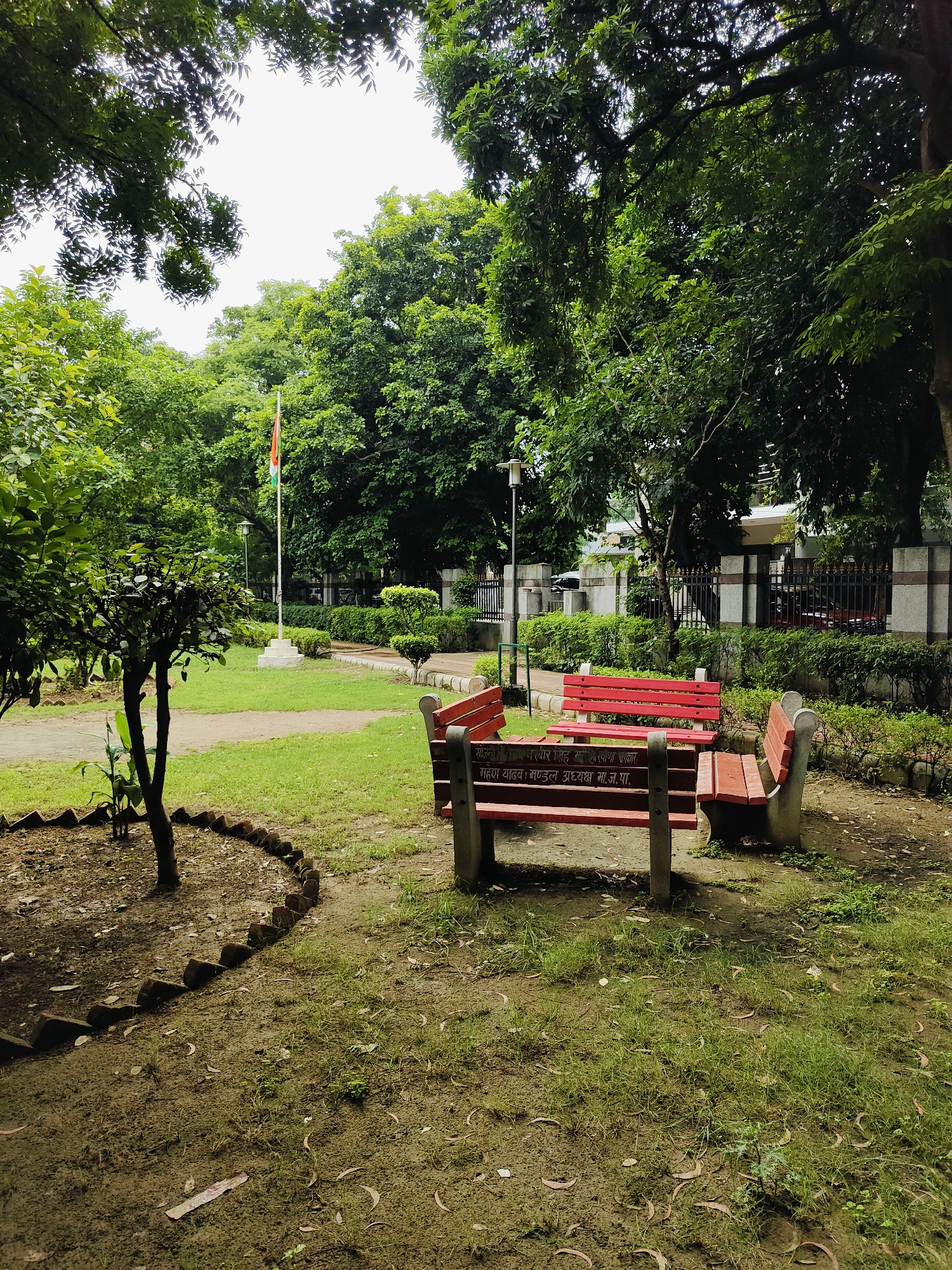 The image depicts an outdoor scene in a park, likely in Gurgaon, India, given the presence of an Indian flag. In the foreground, there are several red benches with stone bases arranged in a semi-circle. Some of the benches have inscriptions in Hindi. A small tree with dense foliage is planted to the left, bordered by a semi-circular arrangement of bricks. The ground is a mix of soil and grass, with fallen leaves scattered about.
In the mid-ground, a grassy area leads to a flagpole flying the Indian flag. Beyond the flag, there is a line of well-maintained hedges and a decorative fence. In the background, there are buildings with stone accents and trees forming a dense canopy. The overall atmosphere suggests a peaceful, public park setting. The lighting indicates it is daytime, with overcast skies casting a soft light on the scene. There are no people visible in the image.