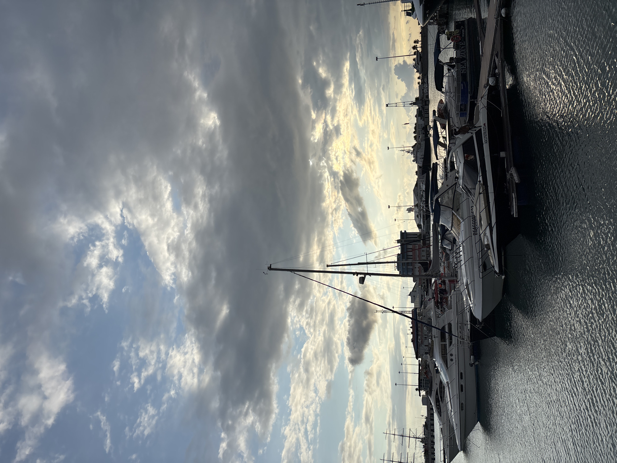 The image shows a marina in Sochi, Russia, with several yachts and boats docked in the water. The sky is filled with dramatic, cloudy skies, with breaks of sunlight illuminating the scene. In the foreground, several motor yachts are lined up, with their hulls reflecting on the rippled water. One of the yachts in the middle ground has a registration number "BE4666RIUS23" visible. Further back, a larger sailboat with multiple masts is docked. The background shows a cityscape with buildings and cranes, indicating a bustling port area. The overall atmosphere is serene yet dynamic due to the interplay of light and clouds.
