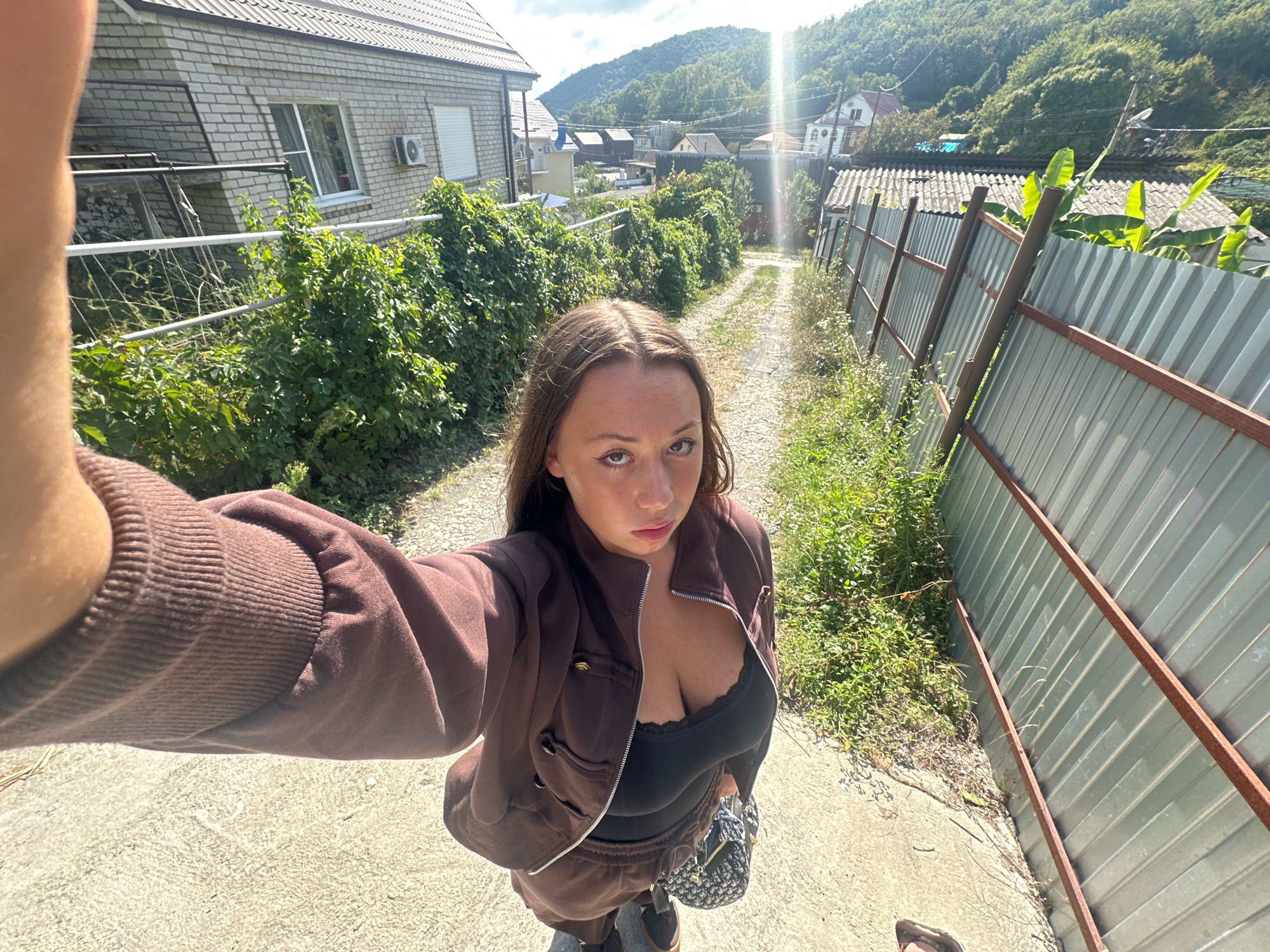 The image shows a young woman standing on a gravel path in what appears to be a rural or suburban area of Lermontovo, Russia. She is wearing a brown tracksuit and a black top. She is holding a camera or phone, taking a selfie. Behind her, there is a metal fence and lush green vegetation. In the background, houses and hills can be seen. The sky is partly cloudy with the sun shining brightly. The overall setting suggests a sunny day in a hilly region.