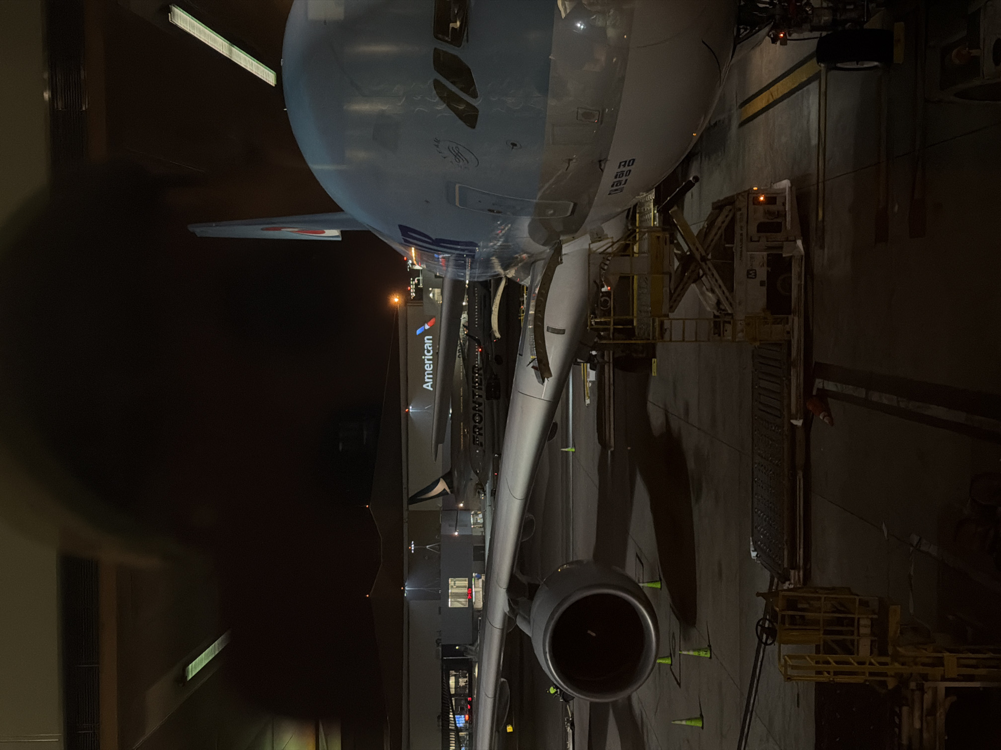 At Los Angeles International Airport, United States of America, an aircraft is parked at a gate, presumably for boarding or disembarkation. Another airplane from American Airlines is parked at a gate next to it. The environment indicates a nighttime setting, likely under the artificial lighting of the airport terminal. A ramp agent or ground crew member may be present at the gate, working near the aircraft.