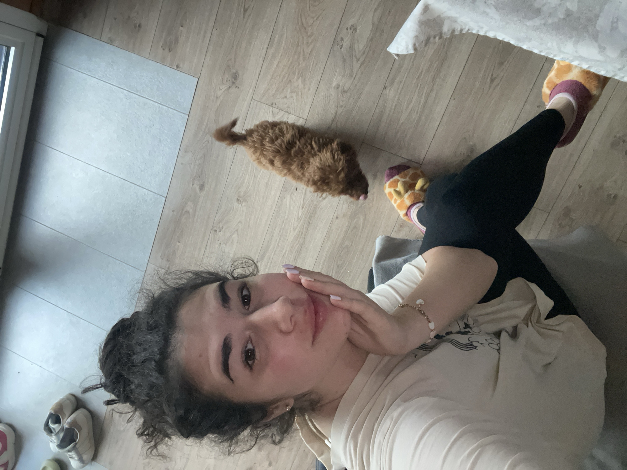 A woman is positioned in the lower-left quadrant of the frame in an indoor setting, possibly an apartment. She is seated and is wearing a light-colored shirt, black leggings, and giraffe-patterned slippers. She is looking at the camera with her hand near her face. Her hair is dark and curly. To the upper-left, there is a window and some shoes on a tiled floor. In the upper half of the image, a small brown dog is lying on a wood-patterned floor, near the woman's feet. A portion of what appears to be a white tablecloth is visible in the upper-right corner. There is no discernible information to indicate that the location is in the city of Sofia, Bulgaria.