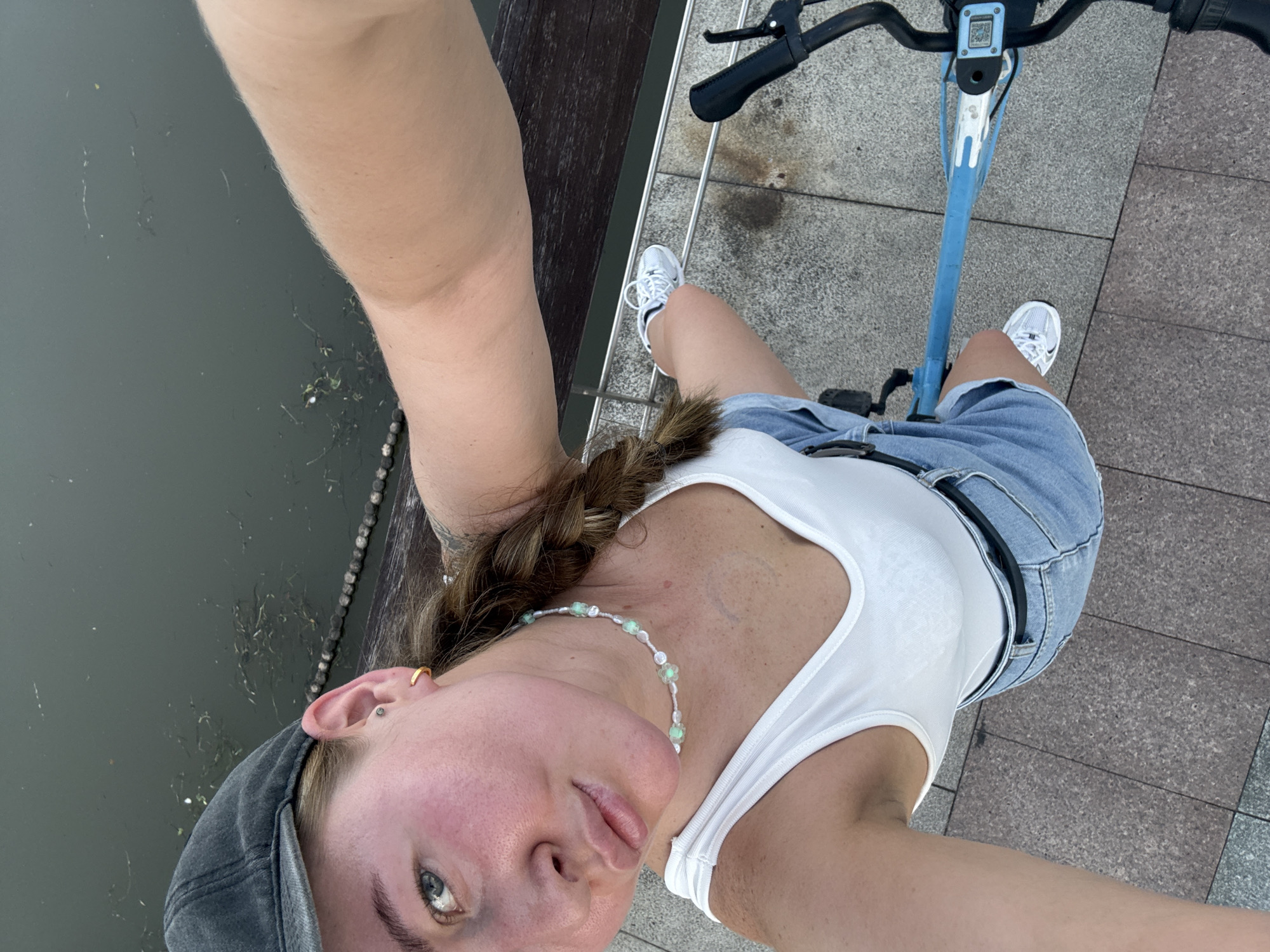 A first-person perspective selfie shows a young woman with a braided ponytail, wearing a white tank top, denim shorts, and a grey baseball cap. She is seated on a blue electric scooter, with her left arm extended, holding the camera. The scooter's handlebars, display screen, and front wheel are visible. She is wearing white athletic shoes.
The background features a tiled walkway and a body of water with some debris. A dark wooden bench is also visible to the left. The lighting suggests it is daytime, and the weather appears to be clear.
Notable details include the woman's jewelry: a beaded necklace with light green beads and floral accents, and a small hoop earring in her left ear. She has a subtle smile and appears relaxed. A QR code is visible on the scooter's display screen.
The scene is set in Yiwu, China.