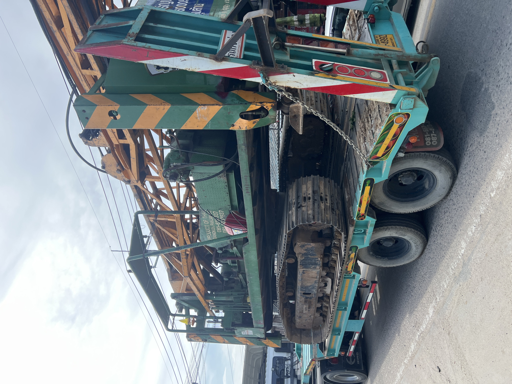 The image shows a large, green and yellow tracked drilling machine being transported on a teal flatbed trailer. The trailer is being towed by a truck, part of which is visible on the left side of the frame. The drilling machine is quite large and appears to be made of metal, with various pipes, cables, and machinery visible. The trailer has multiple wheels and reflective safety markings. The scene is outdoors, with a cloudy sky overhead. The ground appears to be asphalt or a similar road surface. There is some foliage visible in the background. A license plate on the back of the trailer is visible and reads "LAND 51 -4992". There are also some Thai characters visible on the side of the drilling machine and on the trailer. The overall impression is of a heavy-duty industrial vehicle being transported for construction or other heavy works. The context of Ko Pha-ngan District, Thailand is not explicitly indicated by any visible landmarks or signage in the image itself, but it is provided as the location.
