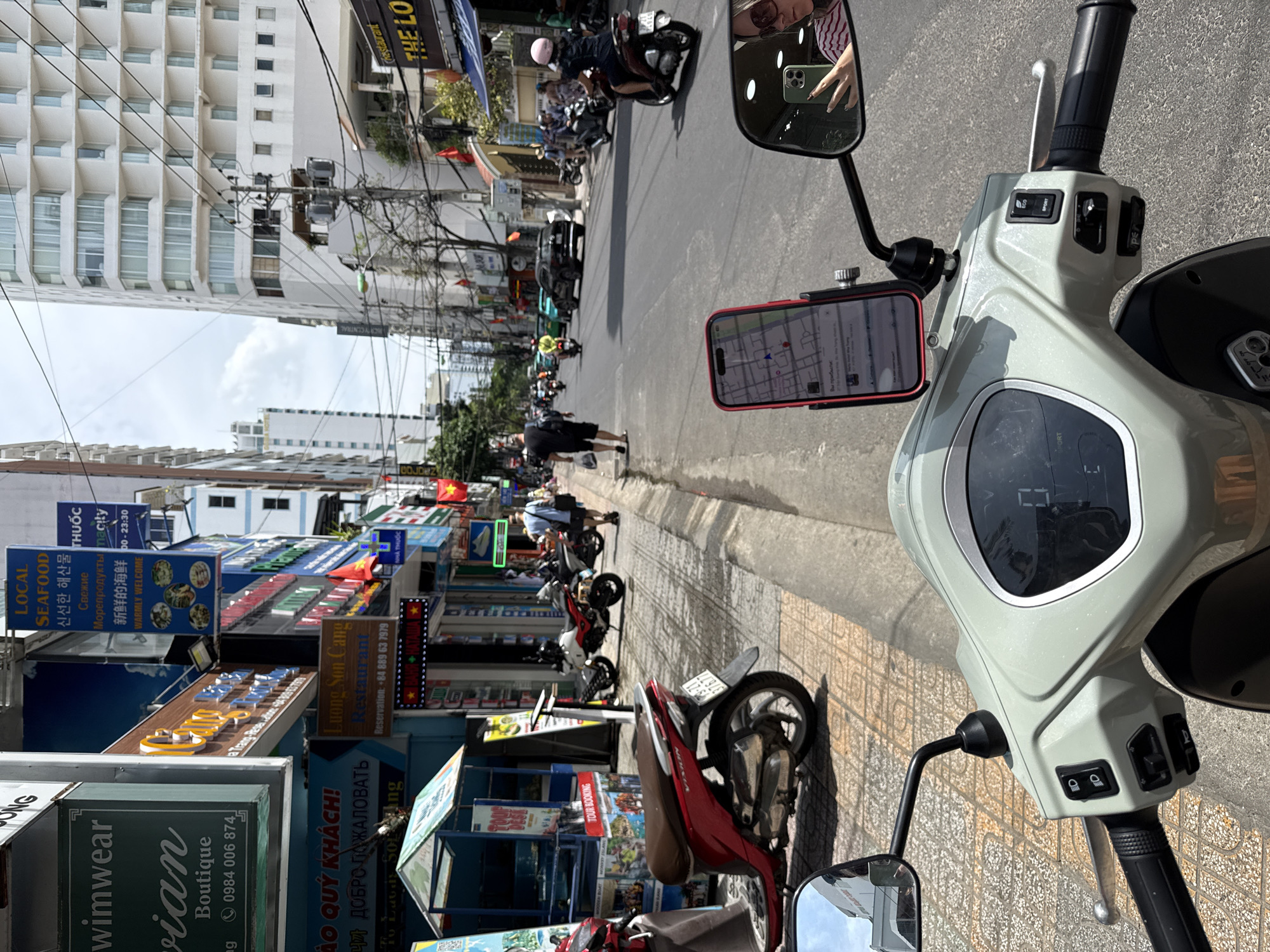 The image is taken from the perspective of a person sitting on a scooter in Nha Trang Ward, Vietnam. The scooter features a phone mount with a navigation app displayed, alongside standard controls and mirrors. A pedestrian and several other motorbikes are moving along the street. Shop signs for businesses such as "Seafood," "Wimwear Boutique," and "Luong Son Cang Restaurant" are visible. In the reflection of the scooter's side mirror, a person is taking a photo with a green smartphone. Taller buildings rise above the shops, with power lines strung overhead. The overall lighting indicates a sunny day.