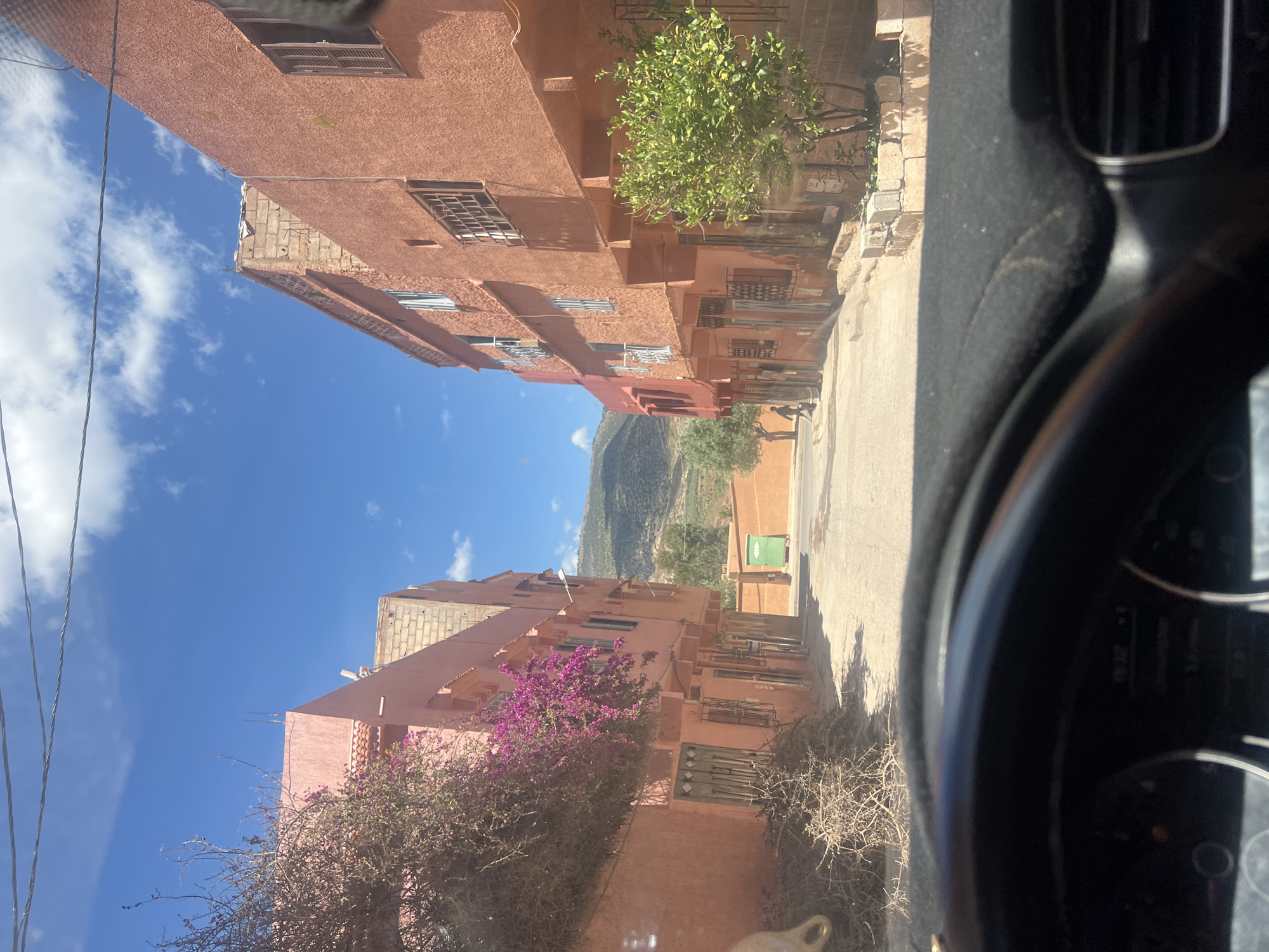 The image, taken from inside a vehicle, shows a street scene in Ouaouizert, Morocco. The street is flanked by peach-colored buildings with barred windows and brickwork. A green tree and a flowering purple bush are visible on either side of the road. A mountain range appears in the distance. The sky is blue with scattered clouds. Part of the car's dashboard and steering wheel are visible in the foreground. Two people are walking in the distance on the right side of the street.