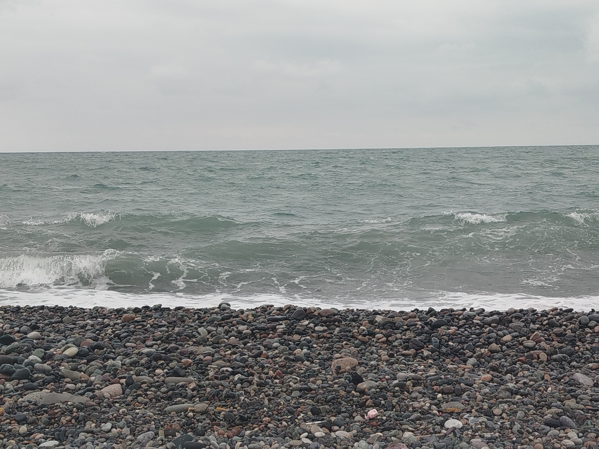 The image depicts a pebble beach with the sea in the background. The sea is choppy with moderate waves breaking on the shore, creating white foam. The sky is overcast and grey, casting a muted light on the scene. The beach is covered with a dense layer of smooth, rounded pebbles of various sizes and colors, ranging from dark grey and black to lighter shades of brown and grey. Some larger stones are visible amongst the smaller pebbles. In the far right of the frame, a small piece of pink plastic trash is visible amongst the pebbles. The scene is devoid of people or any discernible man-made structures. This appears to be a natural coastal landscape, likely in a location such as the Black Sea coast of Batumi, Georgia, known for its pebble beaches.
