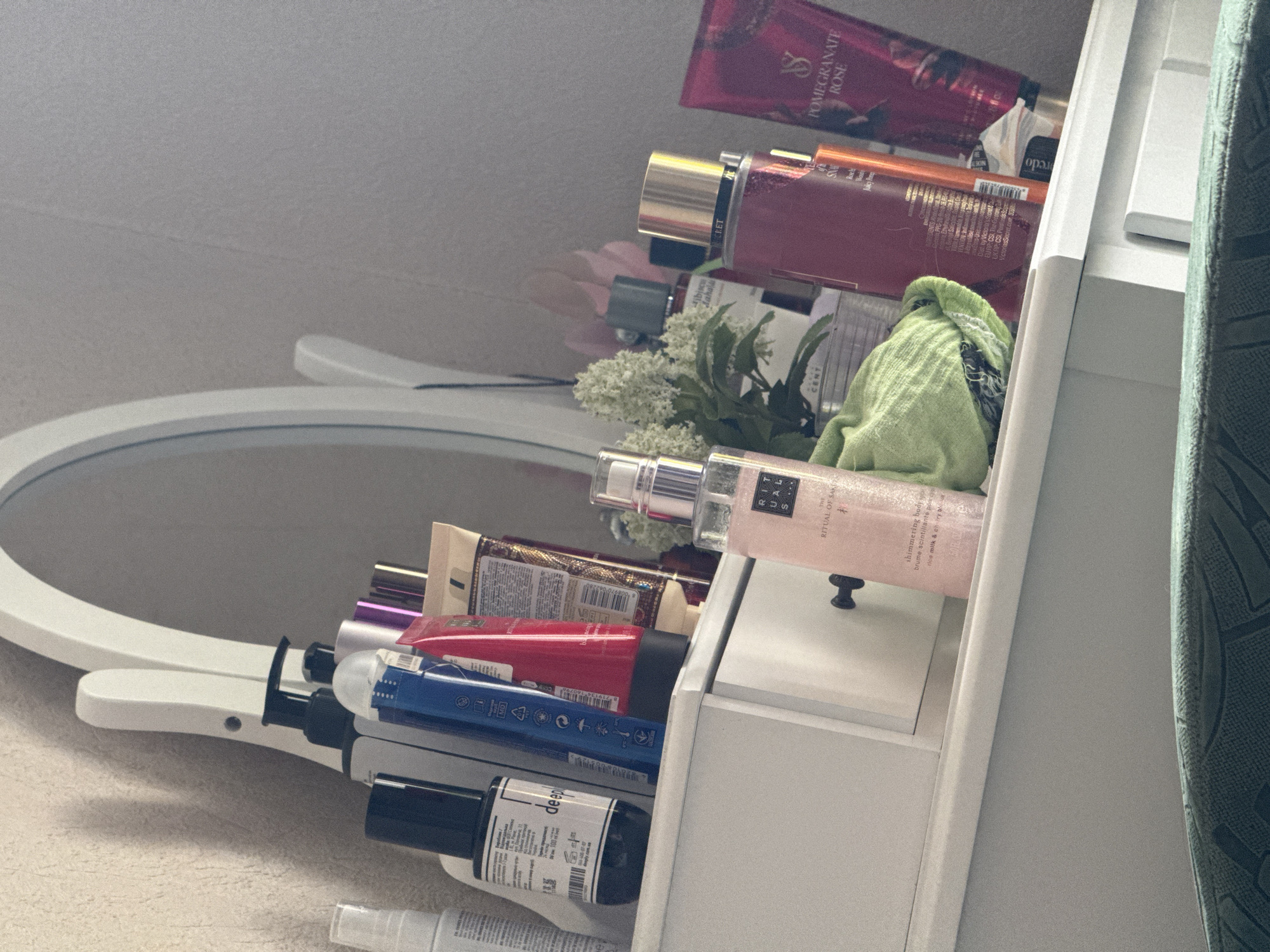 An indoor, eye-level medium shot captures a vanity dresser with various beauty products arranged on its surface. The dresser is white and has a single drawer with a black handle. The products include several bottles and tubes of lotions, perfumes, and other cosmetics. Among them are recognizable brands like Victoria's Secret and Rituals. A white oval mirror with a decorative frame is positioned behind the dresser. The background is a textured light-colored wall. The lighting appears to be natural and somewhat dim.