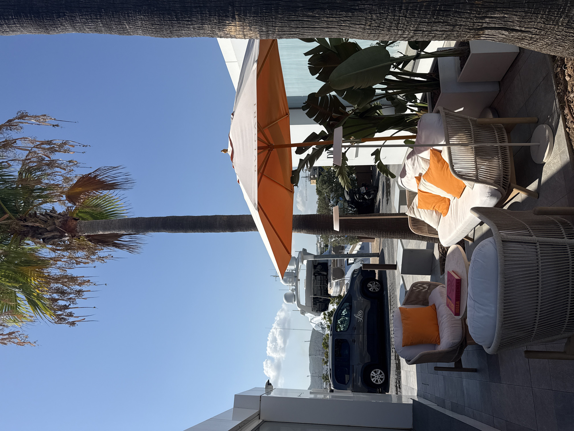 The image depicts an outdoor seating area in the city of Eivissa, Spain, likely a restaurant or lounge, with a backdrop of a marina. In the foreground, there are two wicker armchairs and a wicker sofa with white cushions and bright orange accent pillows. A round marble-topped coffee table sits between the armchairs, holding a pink book. Another small round table is partially visible behind the sofa. An orange umbrella with the word "BULGARI" subtly embossed on it is open, providing shade. Behind the seating area, large potted plants with broad green leaves are situated next to a light blue wall. To the left, a dark grey van with a logo is parked on a cobblestone street. In the background, a large white yacht is docked in the marina, with hills visible in the distance under a clear blue sky with scattered white clouds. Two palm trees frame the scene, one on the right and one in the center, with its fronds extending upwards. The ground is tiled. The scene is bathed in bright sunlight, creating distinct shadows.