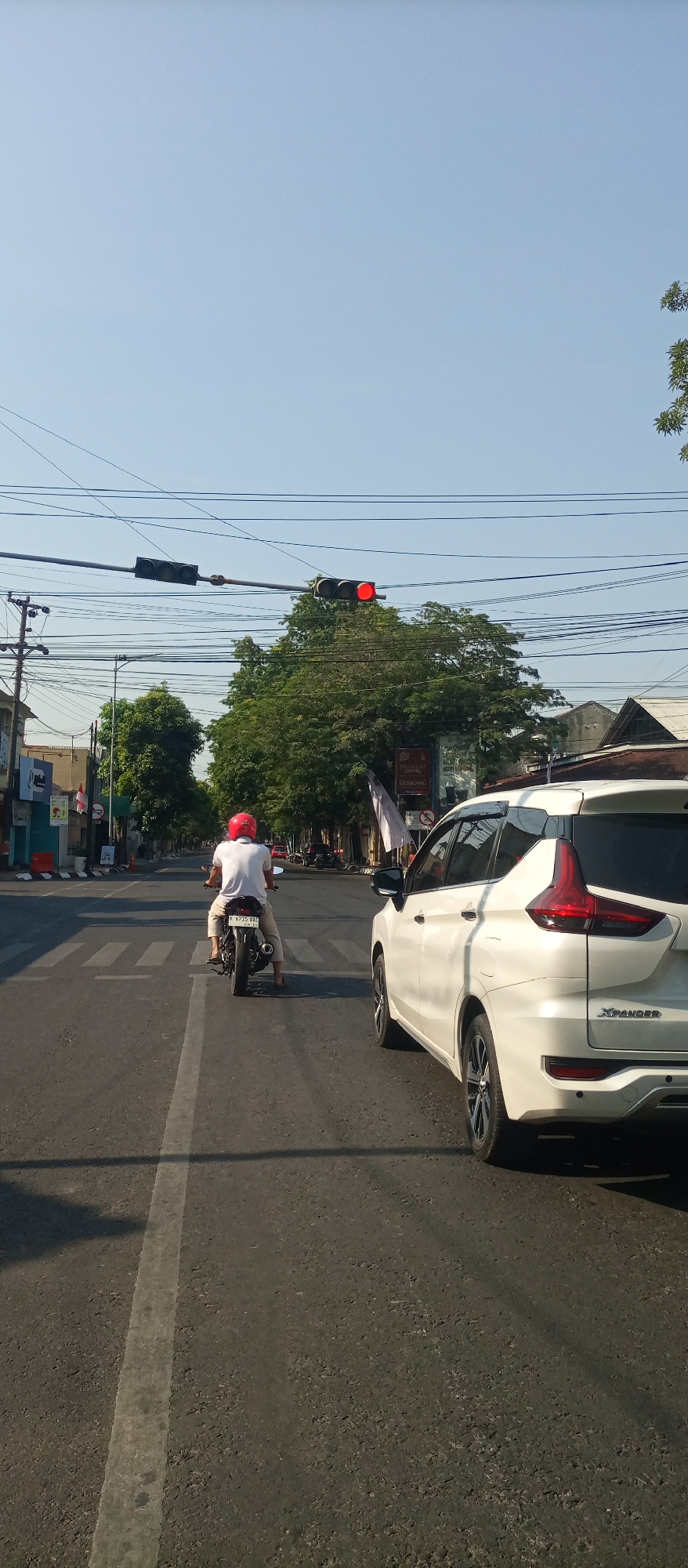 A person wearing a red helmet rides a motorcycle with a white shirt and light-colored pants on a paved road in Panjunan, Indonesia. The motorcycle is moving away from the camera and is positioned in the left lane of two lanes. A white SUV is in the right lane, to the right of the motorcycle. The road has a pedestrian crosswalk marked with white stripes. Overhead, traffic lights hang from wires, with the rightmost light displaying a red signal. Trees line the sides of the road, and buildings are visible in the background. Power lines and utility poles are also present. The sky is clear and blue.