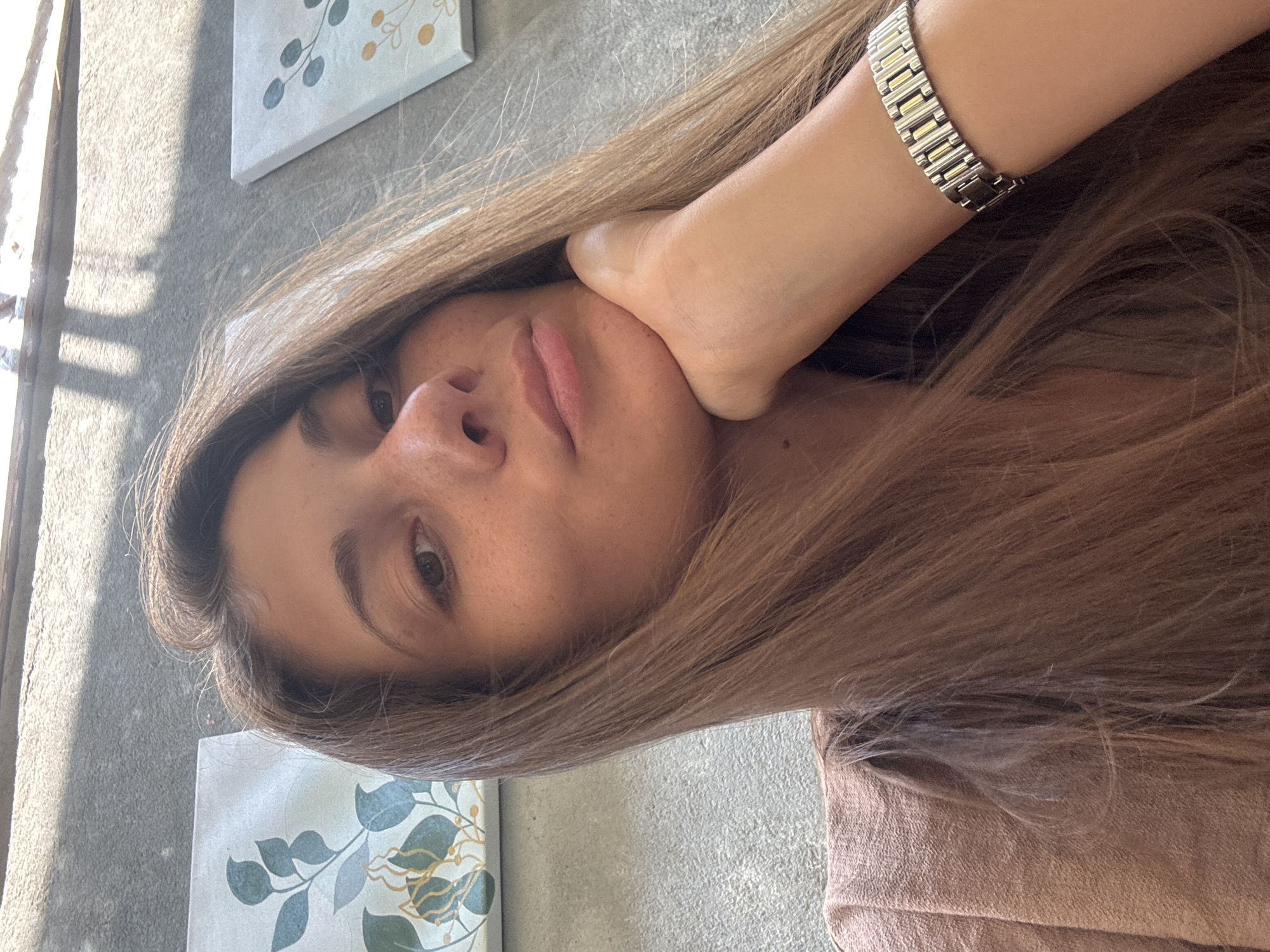 A woman with long brown hair is visible in the image. She is wearing a silver and gold watch on her wrist, and her hand is positioned against her cheek. The background includes a textured grey wall and decorative wall art featuring floral designs. The setting does not give enough details to determine the location within Đà Nẵng, Vietnam.