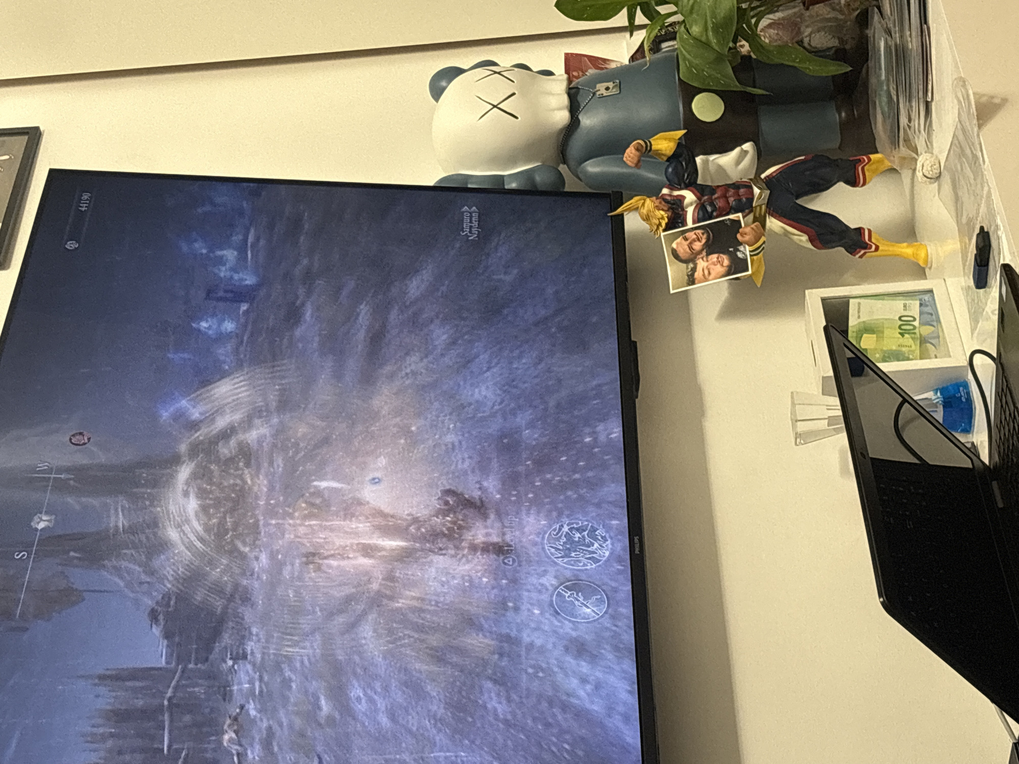 The image shows a Philips television displaying a video game. Above the TV, there is a gray and white figure, a plant, and an action figure of All Might from My Hero Academia. All Might is holding a small photo of two people. To the right of the TV, there is a small white shelf with what appears to be a 100 Euro bill inside. Next to the shelf, there is a black laptop. The location is unknown and cannot be determined from the image.