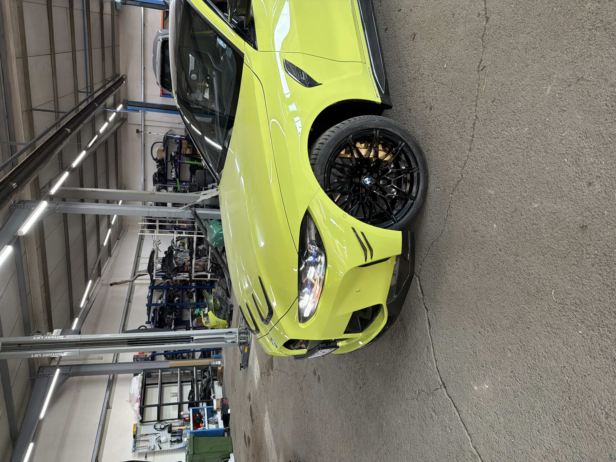 A vibrant, bright yellow-green sports car, likely a BMW M4 or M3, is prominently featured in an indoor automotive workshop or garage. The car is positioned at an angle, showcasing its aggressive front bumper with a visible headlight, intricate black multi-spoke alloy wheel, and gold brake calipers. Fender vents are also visible on the front quarter panel. The vehicle appears to be in pristine, clean condition, reflecting the overhead lighting.
The setting is an industrial space with a concrete floor, which displays some minor cracks and debris. In the background, tall metal shelving units are densely packed with various car components and equipment, indicating a functional parts storage or repair area. Overhead, rows of fluorescent tube lights provide ample illumination. A heavy-duty vehicle lift, clearly labeled "ROTARY LIFT," spans across the mid-ground. No people are visible in the image. The scene suggests the car is either undergoing maintenance, awaiting service, or is stored within this professional automotive environment.
