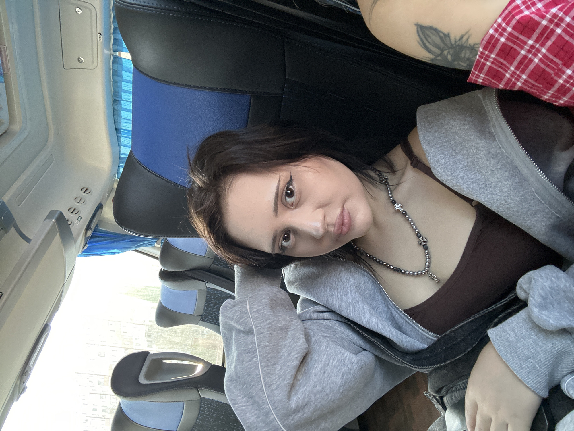 A young woman with dark hair and light skin is sitting inside a bus, possibly in the city of Megion, Russia, given the context. She is wearing a dark brown top, a light grey hoodie, and a necklace with silver crosses. She is looking directly at the camera with a slight pout. A visible tattoo is present on her upper left arm, she also has on cat-eye makeup. The bus seats are upholstered in blue and grey. Outside the window, buildings are visible.