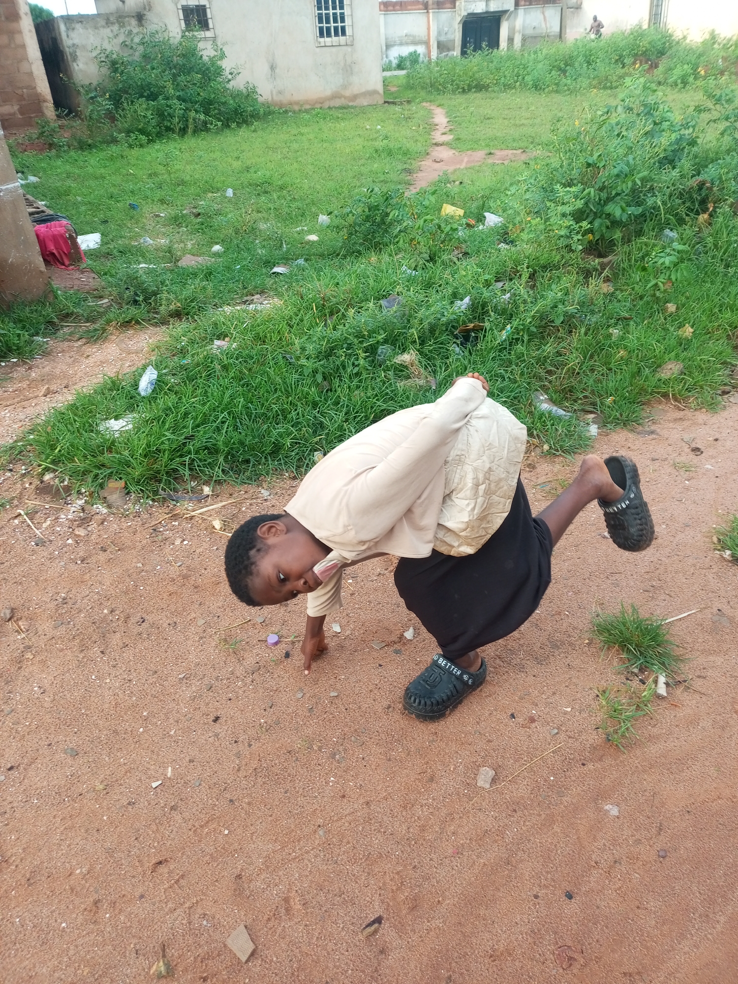 A young Black person, with dark skin and hair, is crouching on sandy ground in front of a grassy area with some scattered debris, including plastic and paper. The person is wearing a light-colored, loose-fitting shirt and a black skirt. One foot is visible, wearing a dark sandal with the word "BETTER" printed on it. The person is looking down and pointing with their right index finger. In the background, there is a simple building with windows and some overgrown vegetation. The overall impression is a casual outdoor scene in a somewhat unkempt environment. The location is identified as Tempure, Nigeria.