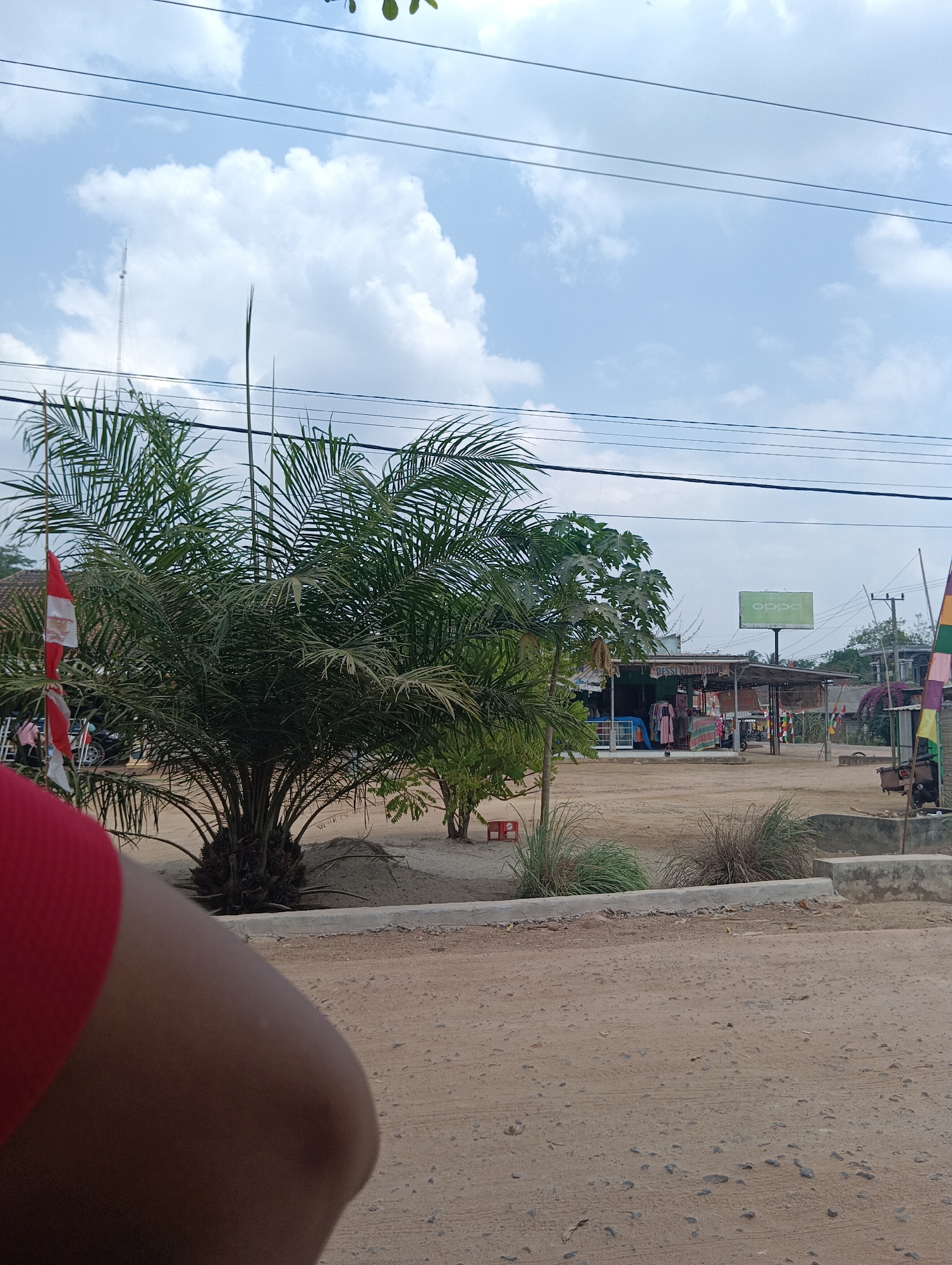This is an outdoor, eye-level, full shot of a dusty, rural road leading to a small roadside market in Binakarsa, Indonesia. In the foreground, the shoulder of a person wearing a red shirt is visible on the left. The road is composed of dirt and small stones. The middle ground features a palm tree with a red and white Indonesian flag attached to its trunk. Behind the palm tree is a small, open-air market with several stalls selling clothing and other items. A large green billboard with the "OPPO" logo is visible behind the market. In the background, utility poles and wires run across the sky, which is partly cloudy. The scene appears to be a typical day in a rural Indonesian town.