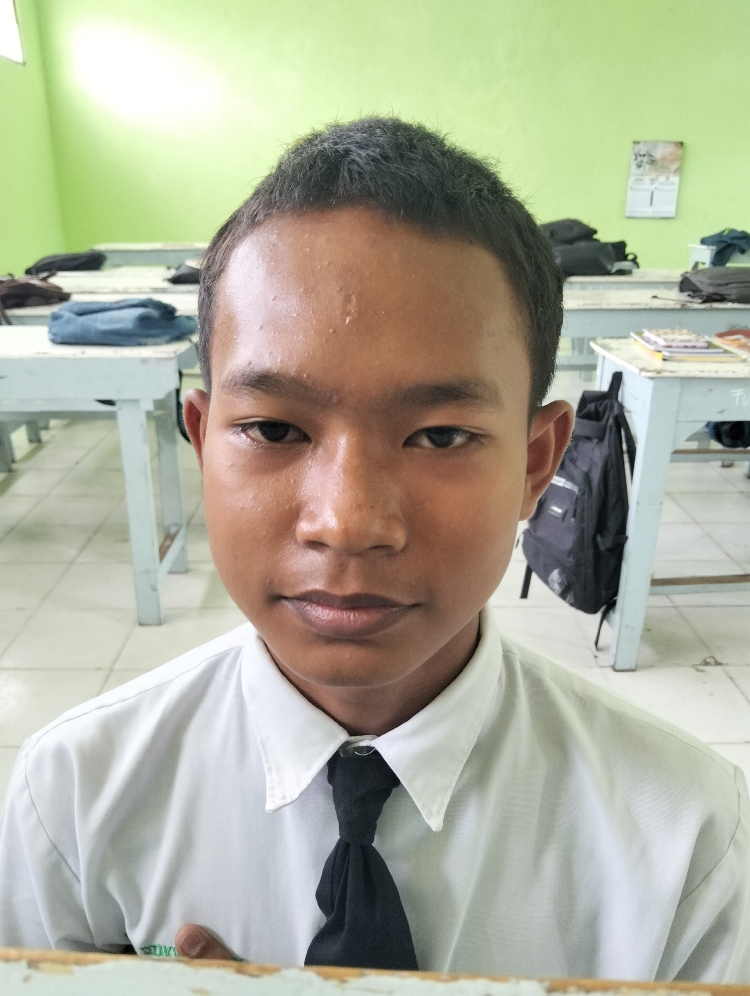 The image shows a close-up portrait of a young male, likely a student, in what appears to be a classroom. He is wearing a white collared shirt and a black tie. His hair is dark and short. His facial expression is neutral. The background reveals rows of empty desks and chairs, some with backpacks or books on them, and a green wall. A calendar is visible on the wall. The lighting suggests an indoor setting, possibly during daytime. The overall scene is consistent with a school environment in Kranji, Indonesia.