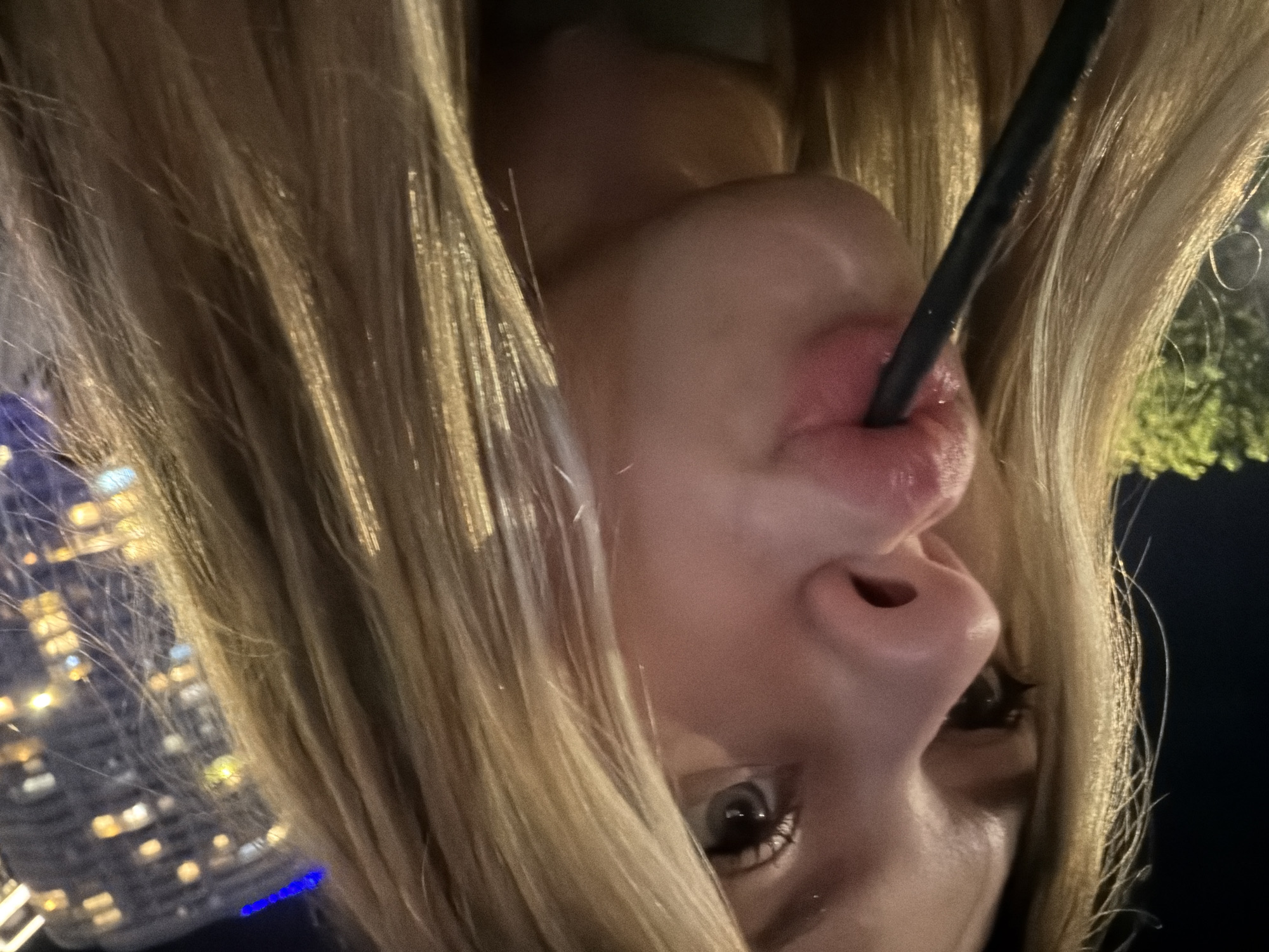A close-up, profile photograph taken at night in Đà Nẵng, Vietnam, captures a young individual with long, straight blonde hair. The subject is positioned on the left side of the frame, angled towards the right, with their light-colored eyes visible. Their prominent nose is in the foreground, and their lips are pursed around a thin, dark-colored straw, indicating they are drinking. The blonde hair is illuminated by artificial light, showing distinct strands and highlights. A dark green garment is partially visible at the bottom of the frame. The background on the immediate left features blurred green foliage under dim light. On the right, a blurry urban landscape at night includes a tall building with multiple illuminated windows, consistent with a city environment. A faint blue light is visible in the upper right background.