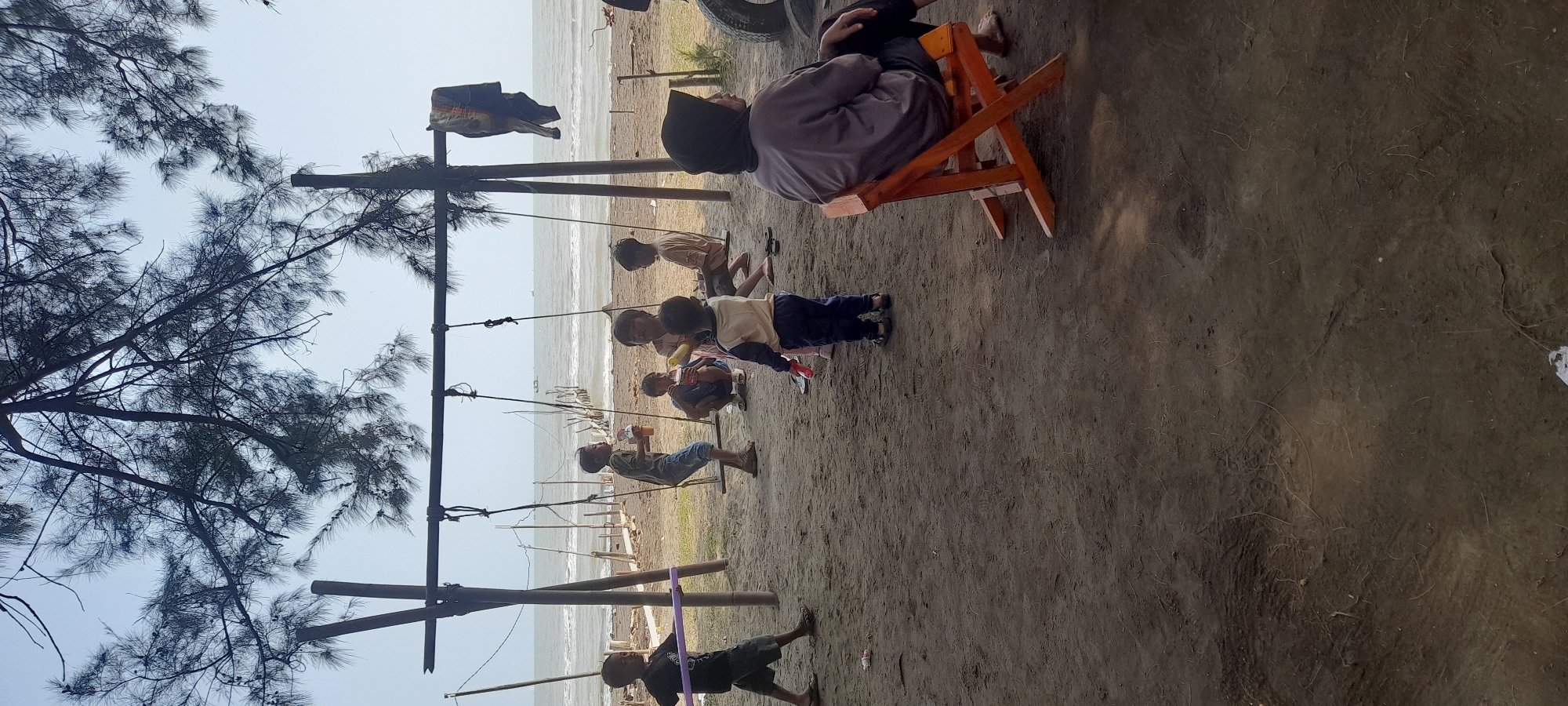 This photograph captures a scene at Nyamplung Sari Beach, Indonesia, on a bright, clear day. A group of children are actively engaged near a rustic swing set constructed from wooden poles and a horizontal beam. One boy, wearing a dark t-shirt and shorts, walks from the left side of the frame holding a purple cylindrical object. Further into the scene, another boy in a green t-shirt and blue shorts stands near a swing, holding a cup. Several other children are also present, some on the swings and others sitting on the sand. To the right, an adult, likely a woman wearing a headscarf and a grey shirt, sits on an orange wooden chair facing away from the camera. The background reveals the calm ocean with a few boats visible in the distance, and a wooden pier extends into the water. The ground is sandy and a few pieces of debris are scattered. The overall atmosphere suggests a leisurely day at the beach.