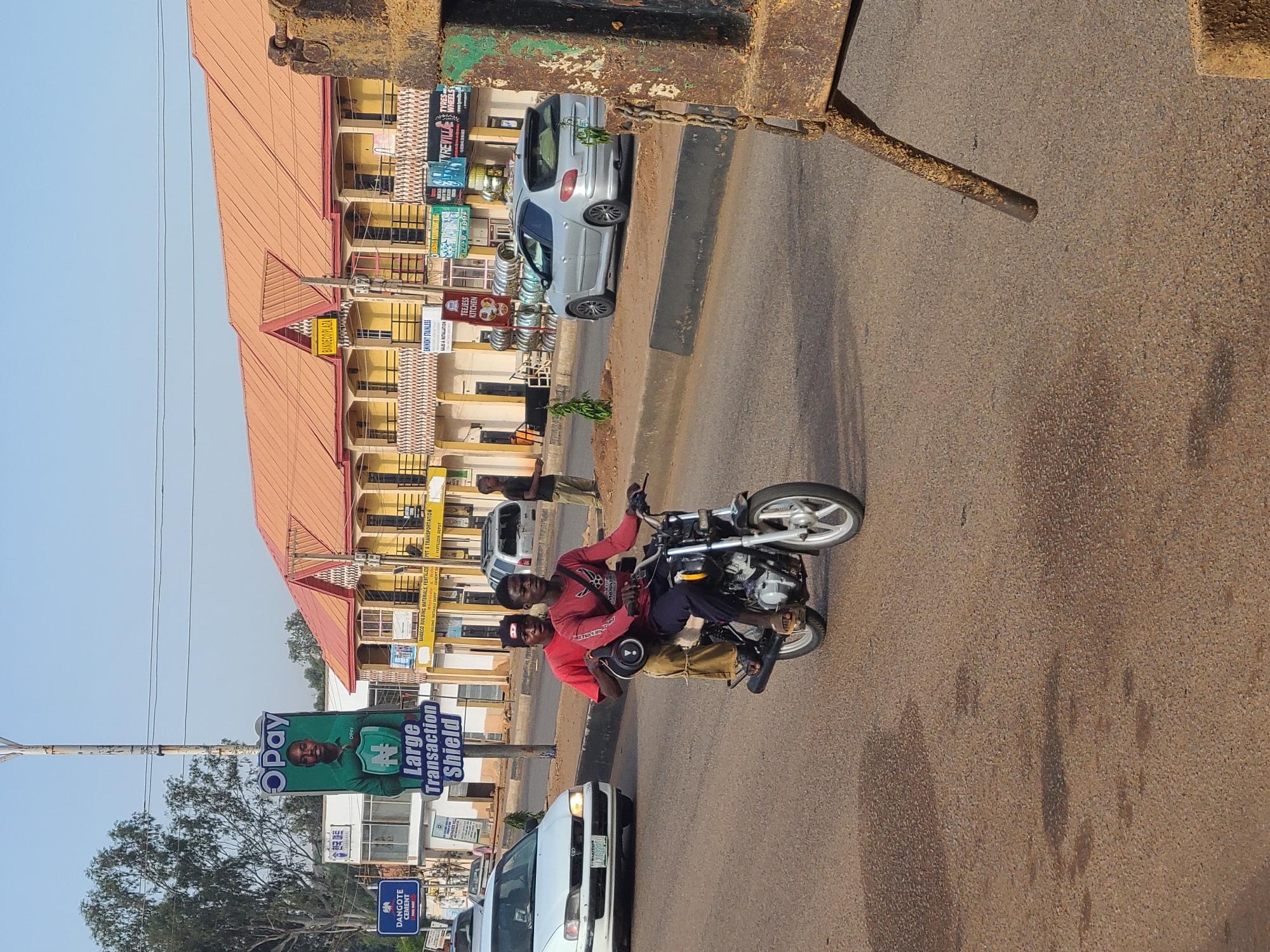 The scene depicts a street in Gura Top, Nigeria, bustling with activity. A motorcycle carrying two individuals, both wearing red shirts, is prominently positioned on the road. The buildings along the street display signs like "BANDECO PLAZA," "EMINENT STAINLESS," "TYREVILLE," and "TEEJESS KITCHEN." A large billboard advertises "Opay" with the slogan "Large Transaction Shield." Other vehicles, including a white car, are visible on the street. In the background, a man in civilian clothing stands on the sidewalk near a parked silver vehicle. The environment appears to be a sunny day with clear skies.