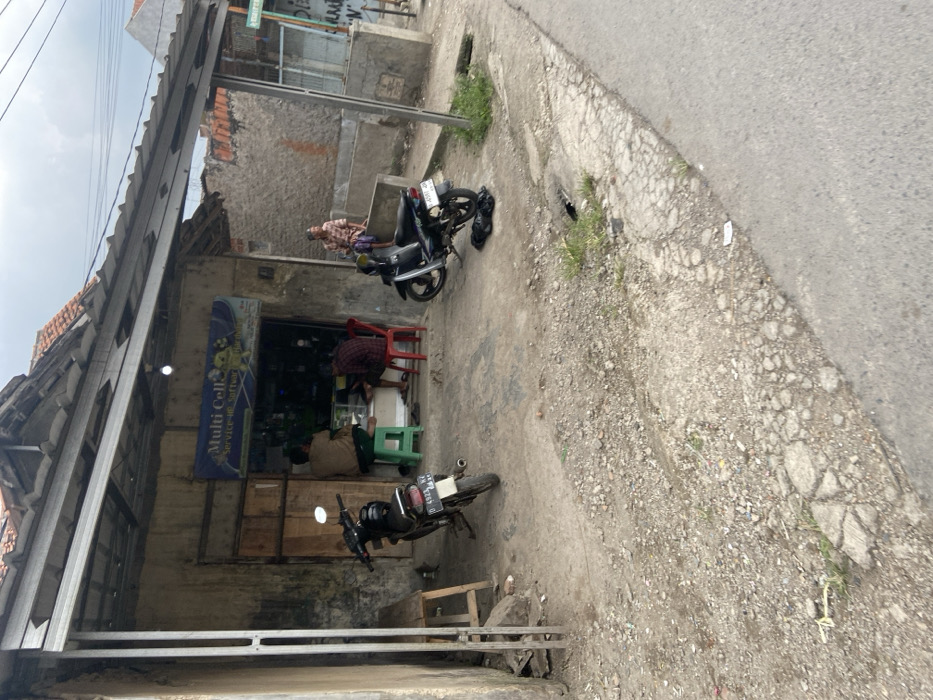 The scene is located in the city of Margaasih, Indonesia at the coordinates -6.965 latitude, 107.542 longitude. There is a small shop on the left side of the image with a sign that reads "Multi Cell Service - Software". Two men are seated inside the shop and a third man stands outside. On the right side of the image, a man stands against a wall with his arms crossed. Two motorbikes are parked in front of the shop. There is a small, wooden, stool in front of the shop. The ground is dirt and gravel. There is a small, paved road on the right side of the image. The sky is overcast. There is a street sign in the distance on the right side of the image. The shop has a metal awning over the entrance. The shop has a metal door and wooden door, both open. The men are wearing casual clothes and are likely customers of the shop. The scene is typical of a small business in a residential area of Indonesia. The scene is peaceful and mundane.