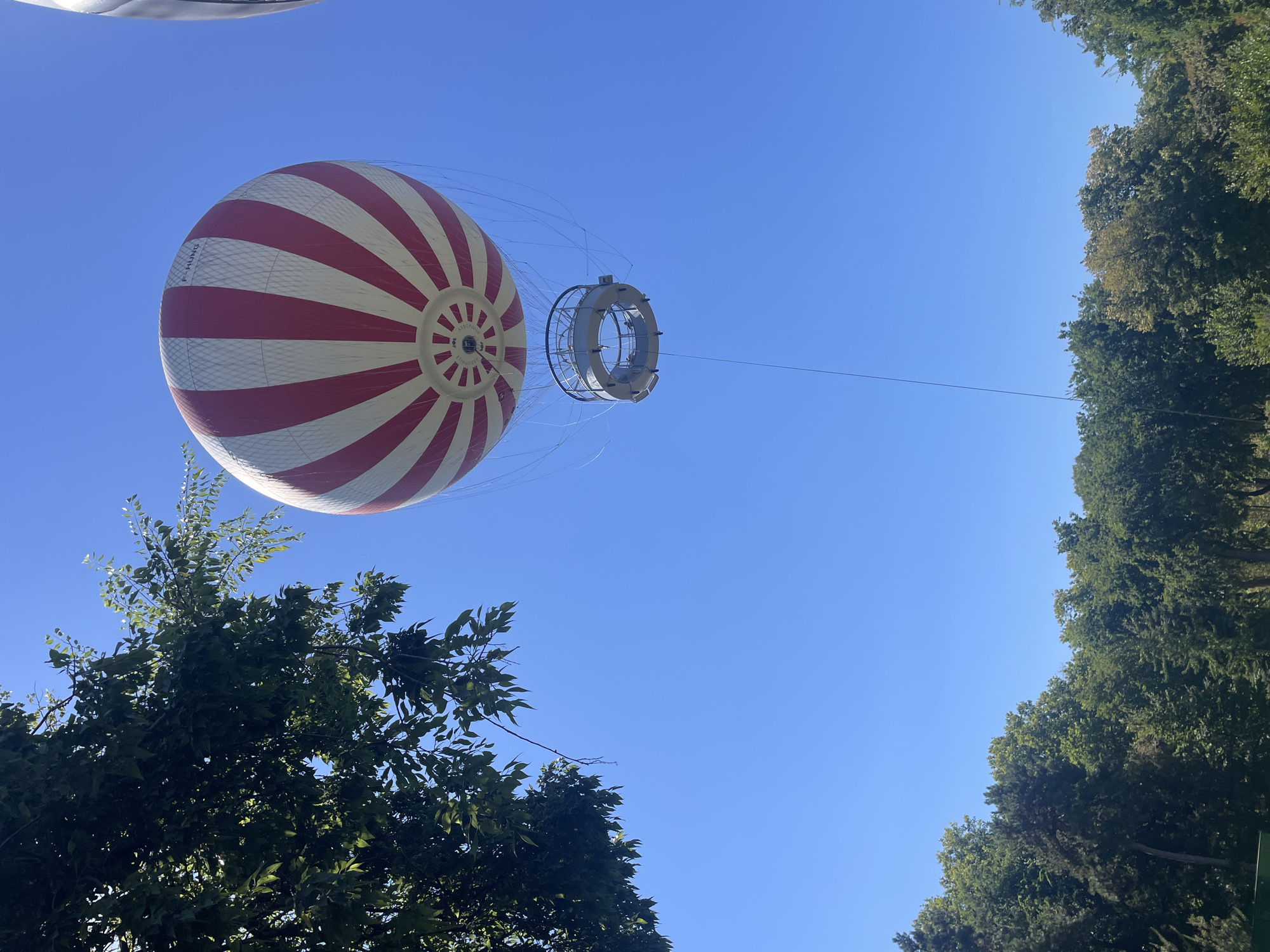 A hot air balloon, with red and white stripes, floats against a clear blue sky. It is tethered to the ground by a thin cable, and a circular gondola hangs beneath it. Trees are visible in the foreground and background. The scene appears to be a park or recreational area, possibly associated with a balloon ride attraction.