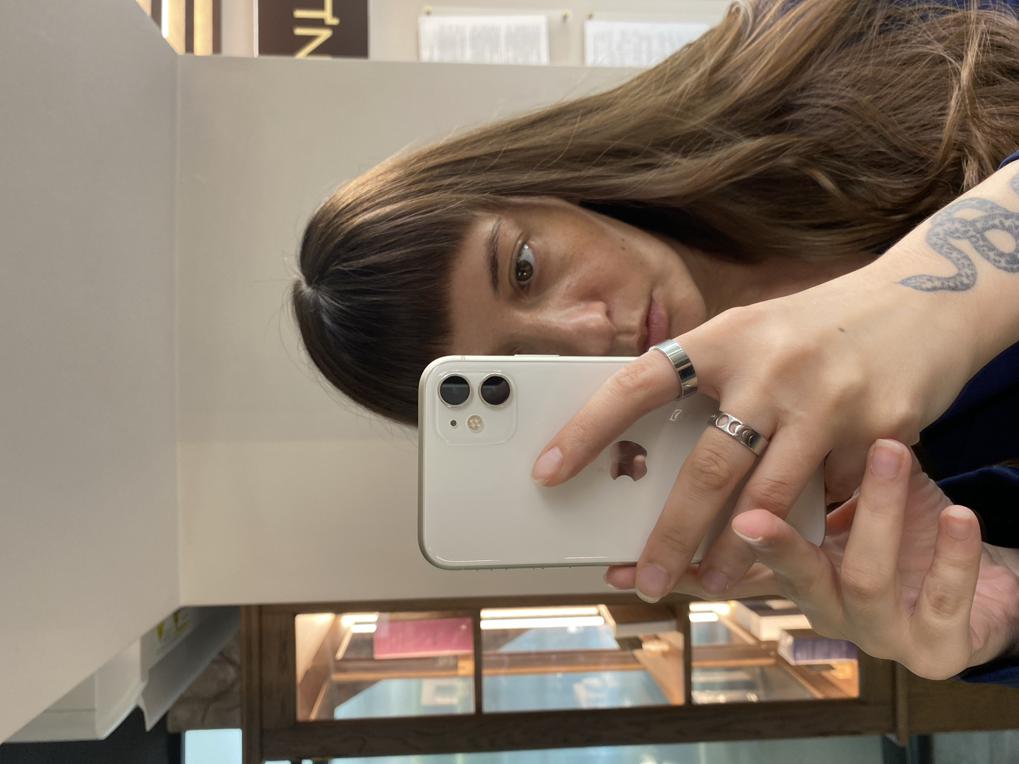 A woman with long, brown, wavy hair and bangs is taking a selfie in a room. She is holding a white iPhone in her left hand, which is angled towards her face. She is wearing a dark blue top and several silver rings on her fingers. One ring on her left index finger has a crescent moon pattern. On her left wrist, a tattoo of a coiled snake is visible. The background shows a wooden display cabinet with shelves containing books and other items. To the right of the woman, a sign with yellow lettering on a brown background is partially visible. The woman's face is turned towards the camera, and she is looking at the phone. The image appears to be taken indoors, possibly in a shop or an office. The location is not Votkinsk, Russia.