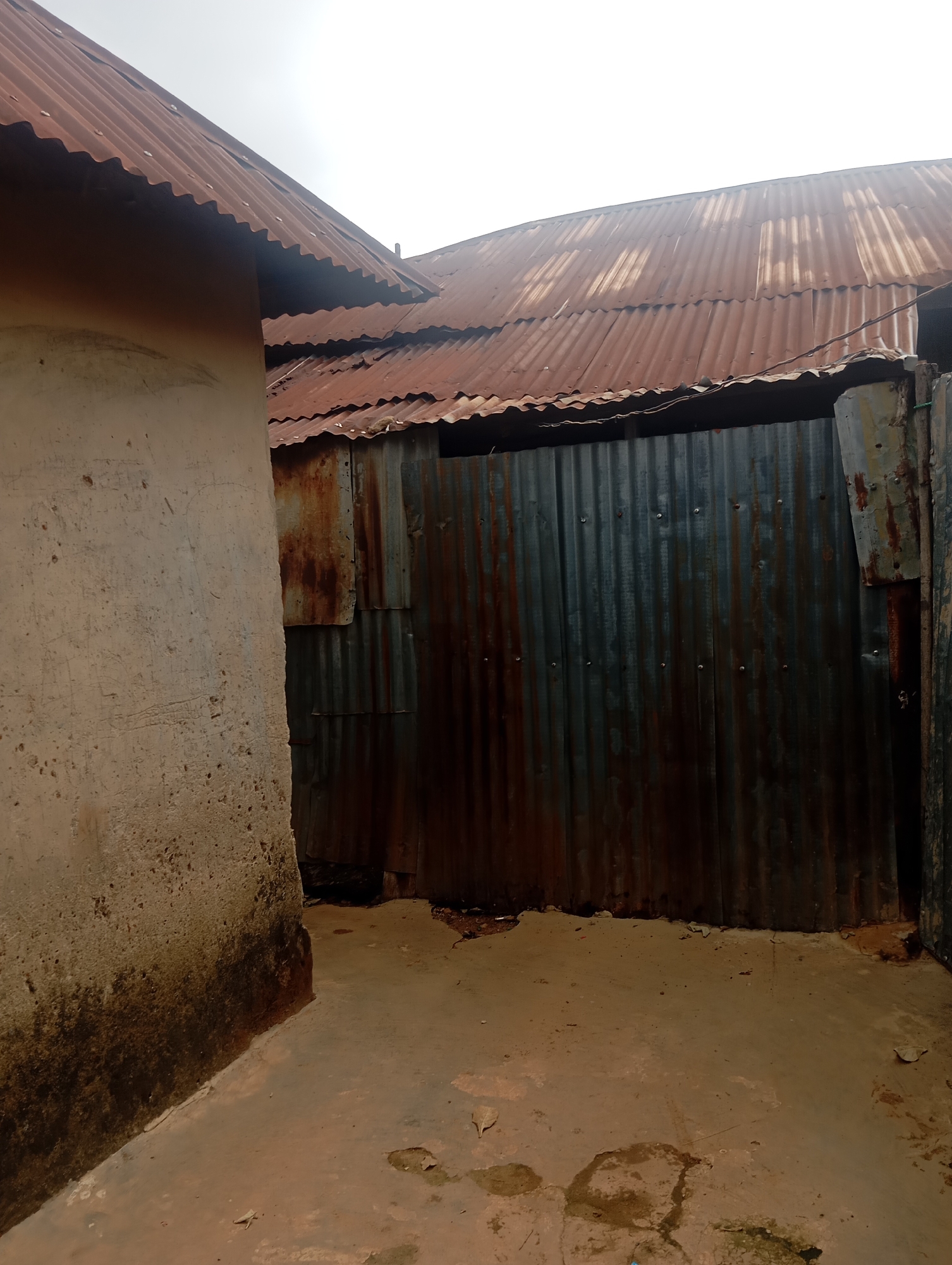 The image depicts an outdoor scene in Dutsen Alhaji, Nigeria. The primary subjects are two adjacent buildings with corrugated iron roofs, showing signs of rust and age. The building on the left has a light-colored, rough-textured wall, stained at the bottom. The building on the right is largely obscured by a wall made of vertically corrugated, rusty metal sheeting. The ground is a mixture of concrete and earth, with some puddles and debris.
The scene appears to be a residential or utility area, possibly a courtyard or alleyway between structures. The overall impression is one of a rural or semi-urban environment, with a sense of wear and tear on the materials.
Notable details include the weathered appearance of the roofing and wall materials, suggesting exposure to the elements over time. The lighting suggests it is daytime, though the sky is overcast, contributing to a muted and somewhat somber atmosphere. There are no people or animals visible in the frame, and no discernible activity is taking place. The location cues are limited to the architectural style and materials common in many parts of Nigeria. There is no visible text in the image.