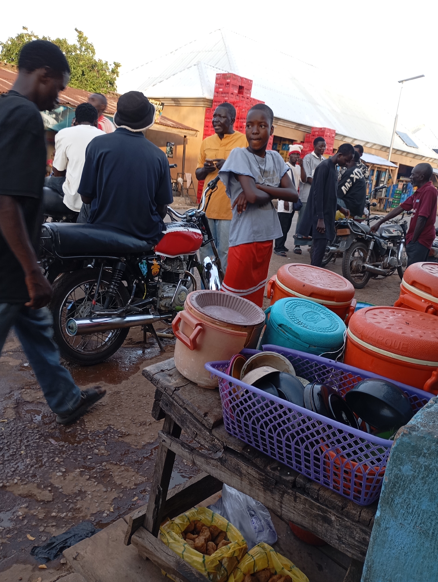 In Shendam, Nigeria, a group of people are gathered in an outdoor marketplace. A young boy in a grey t-shirt and red shorts stands with his arms crossed, looking to his right. Next to him, a man in an orange shirt holds a phone. Several motorcycles are parked in the background. In the foreground, a collection of orange and blue coolers are displayed on a wooden table, alongside a purple basket filled with bowls and lids. Bags of fried food are also visible. The ground appears wet and muddy. The scene is lit by natural daylight.