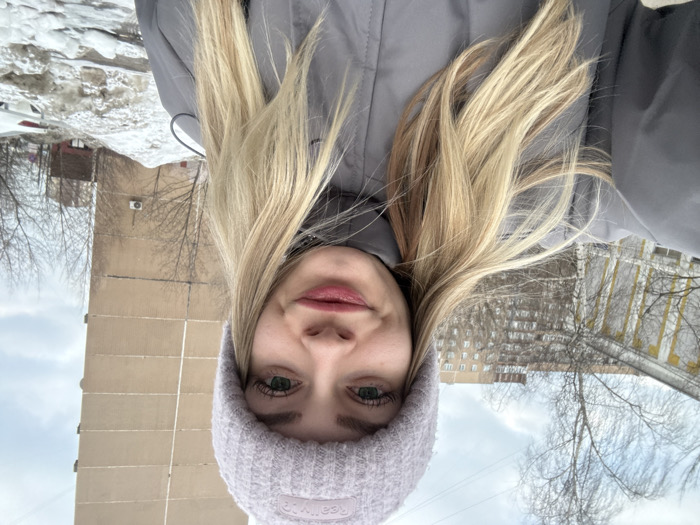 A young adult female is depicted in a close-up, chest-up selfie, positioned outdoors against an urban backdrop on what appears to be a cloudy winter or early spring day. The subject has fair skin, long blonde hair, and green eyes. She is wearing a light lavender knitted beanie with "Really?" inscribed on it and a grey puffer jacket with a high collar. Her lips exhibit light reddish-tinted makeup, and she displays a slight smile. The camera is held at arm's length, suggesting a self-portrait.
The background reveals a street scene with architectural elements and vegetation. To the subject's left, a portion of an older, light-yellow or cream-colored building with visible window frames and stucco details is present. Further behind this structure and centrally located in the mid-ground, a multi-story residential building with a lighter, possibly beige, facade is visible through bare deciduous tree branches. Dominating the right side of the background is a tall, multi-story beige-colored panel apartment building, displaying prominent vertical seams indicative of its construction, with a white air conditioning unit mounted on its facade at a lower level. In the immediate foreground on the right, a substantial pile of dirty, melting snow and ice is visible, consistent with cold weather conditions. The sky above is overcast.
This scene is located in the city of Reutov, Russia. The older, light-yellow building on the left is identifiable as the Children's Art School (Detskaya Shkola Iskusstv) at Pervomayskaya Street, 13. The prominent beige panel building on the right is the residential building located at Pervomayskaya Street, 48. The photograph appears to be taken from an outdoor public area, such as a sidewalk or open space, looking approximately southeast towards these specific landmarks.