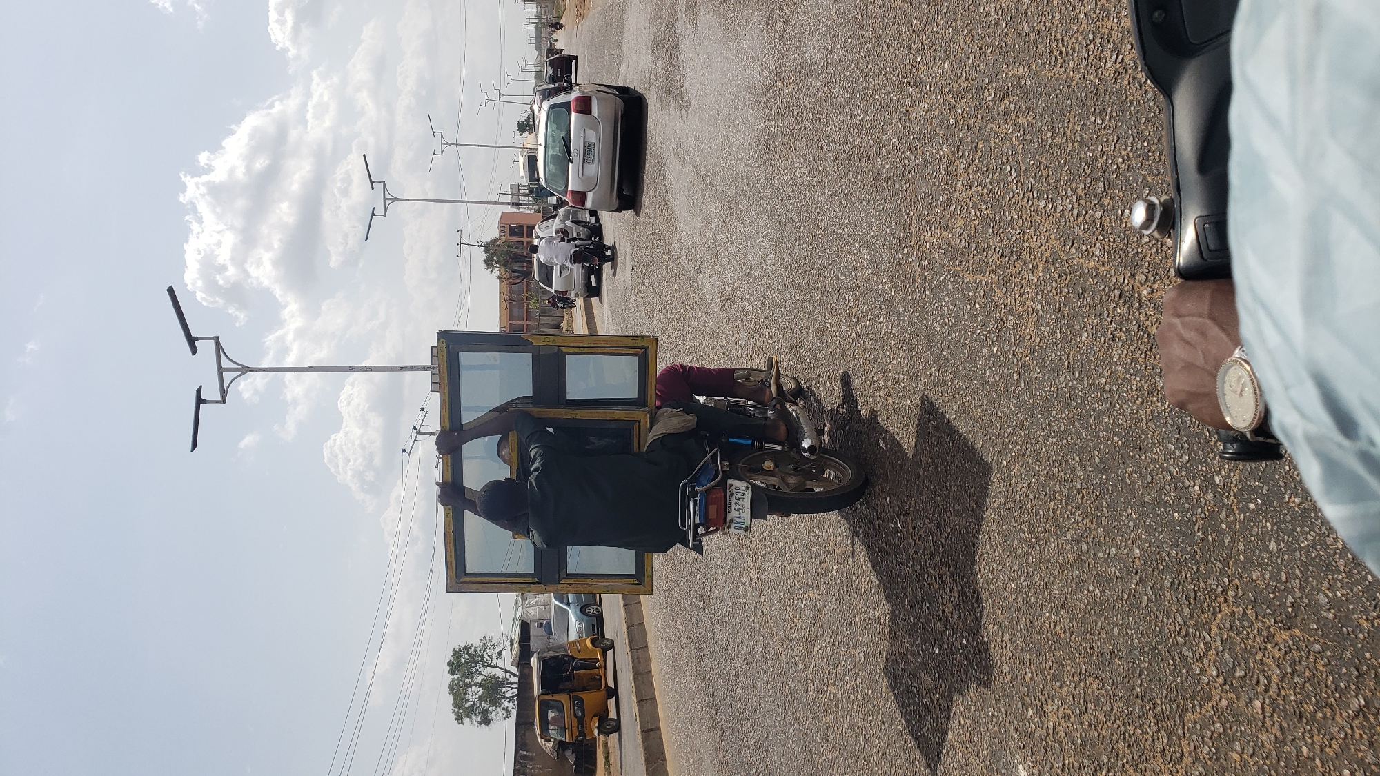 In Hayin Banki, Nigeria, a man is riding a motorcycle on a paved road, precariously carrying a large, rectangular window frame. The frame is held aloft by the man and the motorcycle rider, posing a safety hazard. Other vehicles, including a white car and a yellow auto-rickshaw, are visible on the road. Streetlights are seen along the roadside, and the sky is partly cloudy.