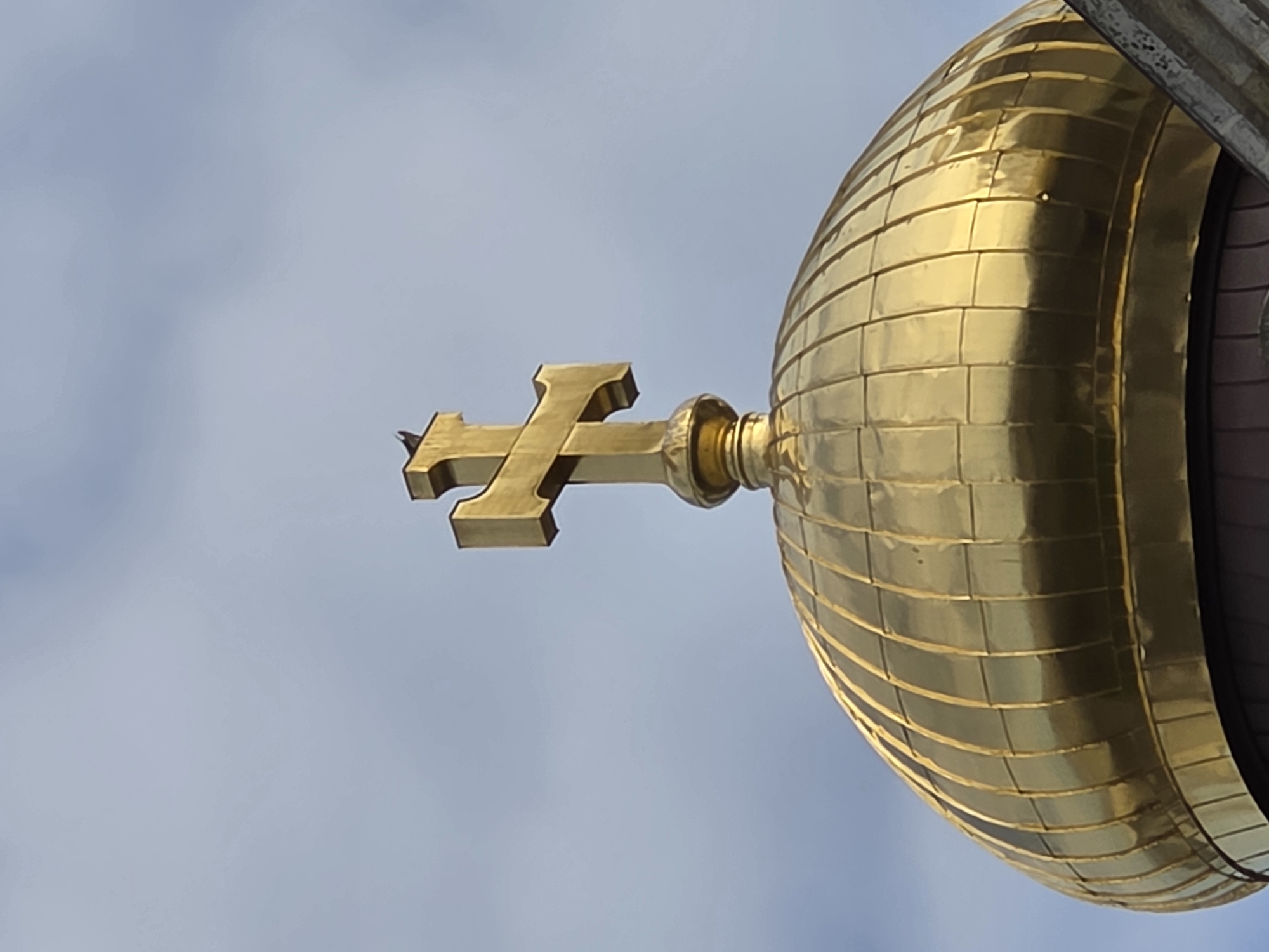 A close-up shot depicts a golden Orthodox cross atop a golden dome against a cloudy sky. The dome has a tiled or paneled surface, and the cross is positioned to the left of the dome, obscuring the view of the sky. The scene is most likely in Varna, Bulgaria, given the architectural style of the dome and cross. There is a bird perched on the left side of the cross.