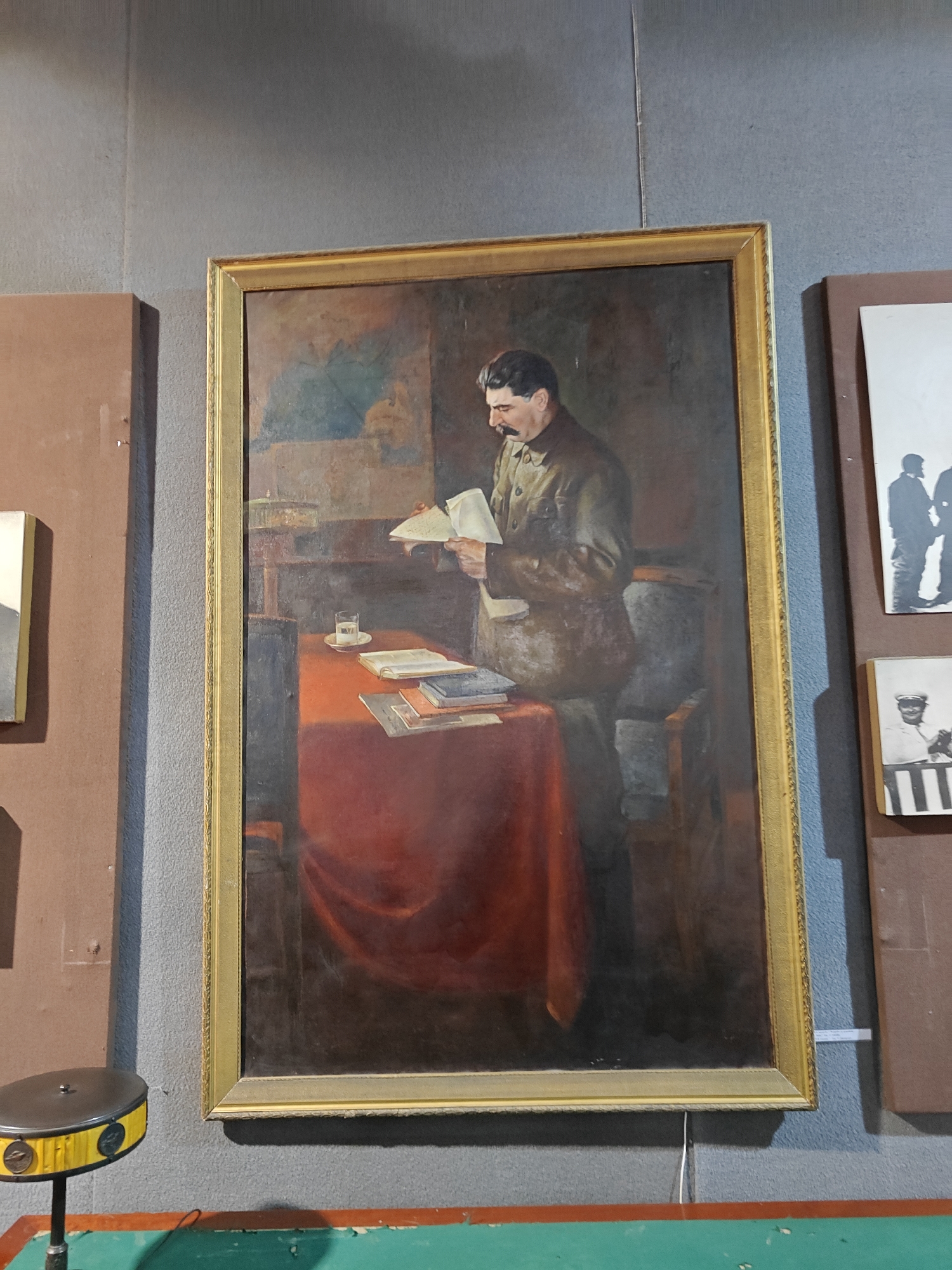 A medium-sized, framed painting hangs on a gray wall in what appears to be a gallery or museum, located in the city of Unknown, Unknown. The painting depicts a man with a mustache, wearing a dark green or brown military-style jacket, standing and reading a document. In front of him is a table covered with a red tablecloth, on which are various books, documents, and a glass of liquid on a saucer. Behind the man is a muted background, possibly depicting a room or interior setting. To the left and right of the painting are sections of brown panels with additional artwork or pictures mounted on them. A green surface is visible at the bottom of the image, with a yellow and black object sitting atop it on the left-hand side. A thin white wire or string hangs down from the bottom of the painting's frame.