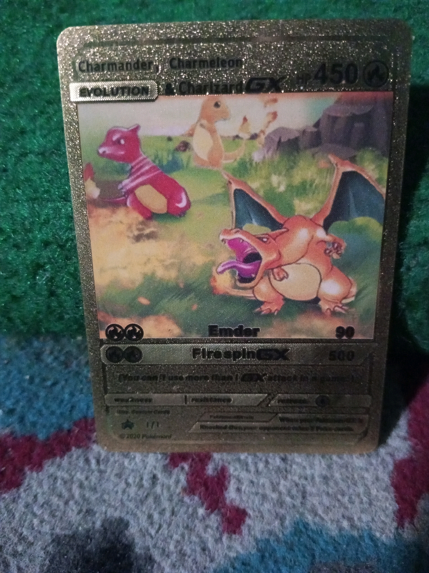 A gold Pokemon card depicting Charizard is laid on a textured surface. The card features the evolution line from Charmander to Charmeleon and Charizard. Charizard, the final evolution, is prominently displayed with its mouth open and tongue out. The card has text, including "EVOLUTION," "Charmander, Charmeleon, & Charizard GX," "Emder," and "Fire spin GX." Below this, there are smaller text elements that are difficult to read due to the image resolution. The background consists of a green speckled surface, partially covered by a blue and red patterned textile. The image is a close-up of the card and its immediate surroundings. There is no indication of the city of Langlang, Indonesia.