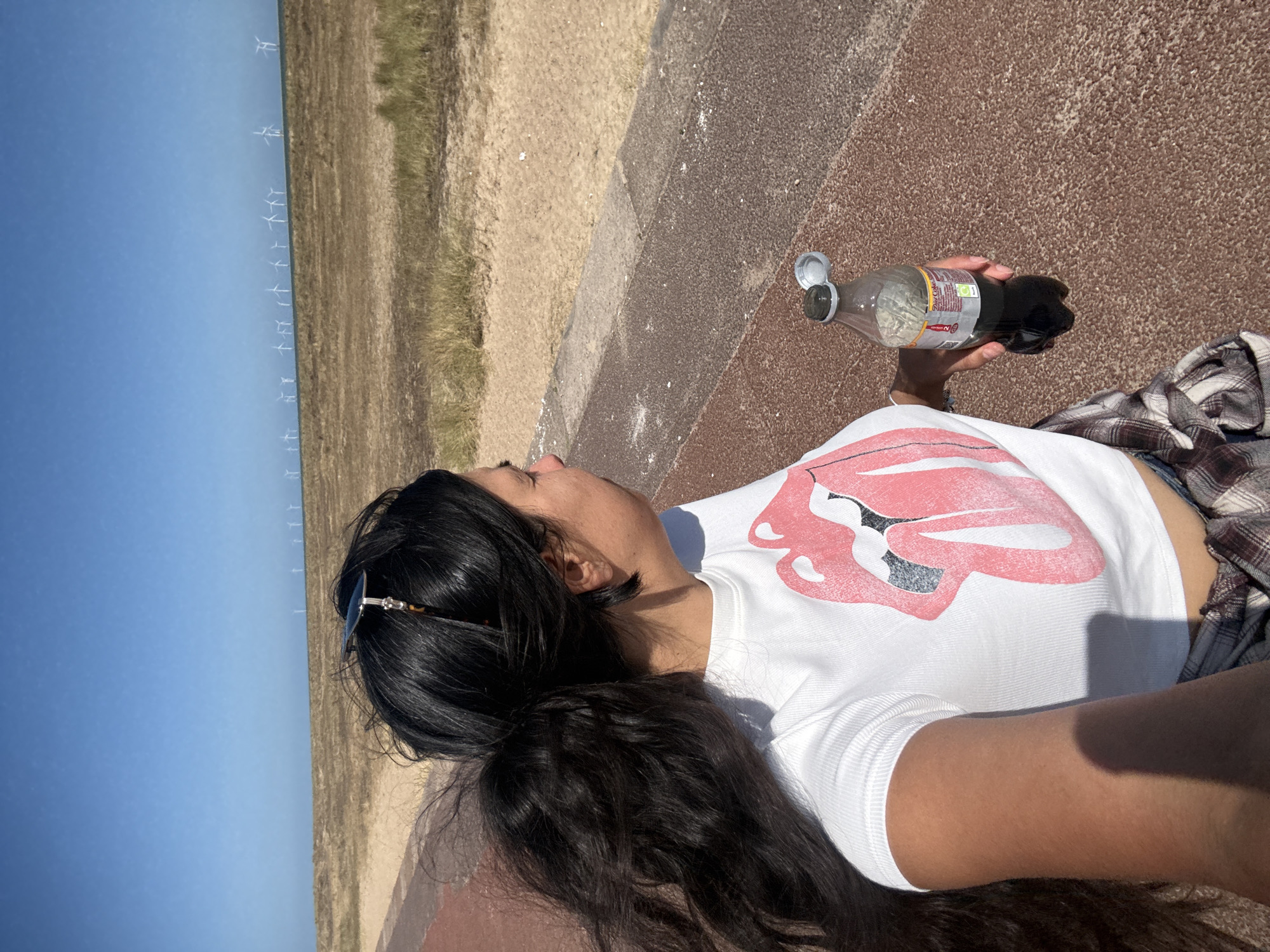 A young woman with long, dark, wavy hair tied in a ponytail stands facing to the right. She is wearing a white cropped t-shirt with a pink Rolling Stones tongue logo on the front, and a plaid shirt tied around her waist. She is holding an open bottle of dark liquid, possibly soda, in her right hand. She is standing on a reddish-brown paved path, with a sandy area and sparse vegetation behind her. In the distance, a line of wind turbines stretches across the horizon under a clear blue sky. The location is the Great Yarmouth seafront, UK.