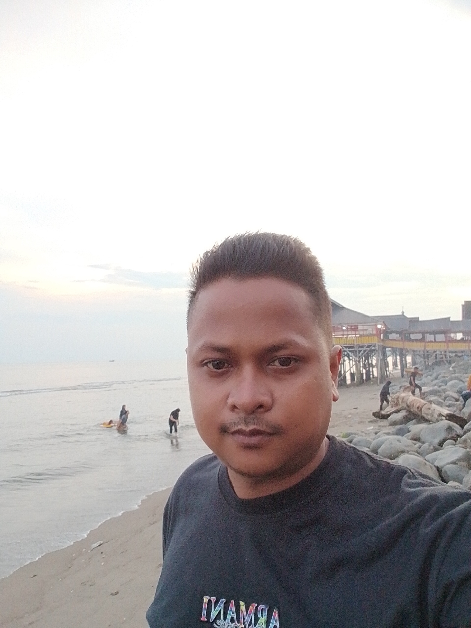 A selfie photograph captures a man in the foreground at a beach in Lhokseumawe, Indonesia. He has dark, short hair styled upwards, a light mustache, and is looking directly at the camera with a neutral expression. He is wearing a dark t-shirt with text on the front that is mirrored, appearing to spell "ARMANI".
Behind him, the scene depicts a calm beach and ocean under a softly lit, overcast sky, suggesting either late afternoon or early evening. Gentle waves wash onto the sandy shore. In the shallow waters to the left, two individuals are visible, one seemingly on a yellow float. Another person stands in the water closer to the right.
Further back on the right side of the beach, there's a rocky area with a large piece of driftwood or a log. Several other people are present on this part of the beach, some near a wooden structure built on stilts over the water, resembling a pier or traditional stilt buildings. The distant sky is mostly white and light grey, indicating soft, diffused light conditions.