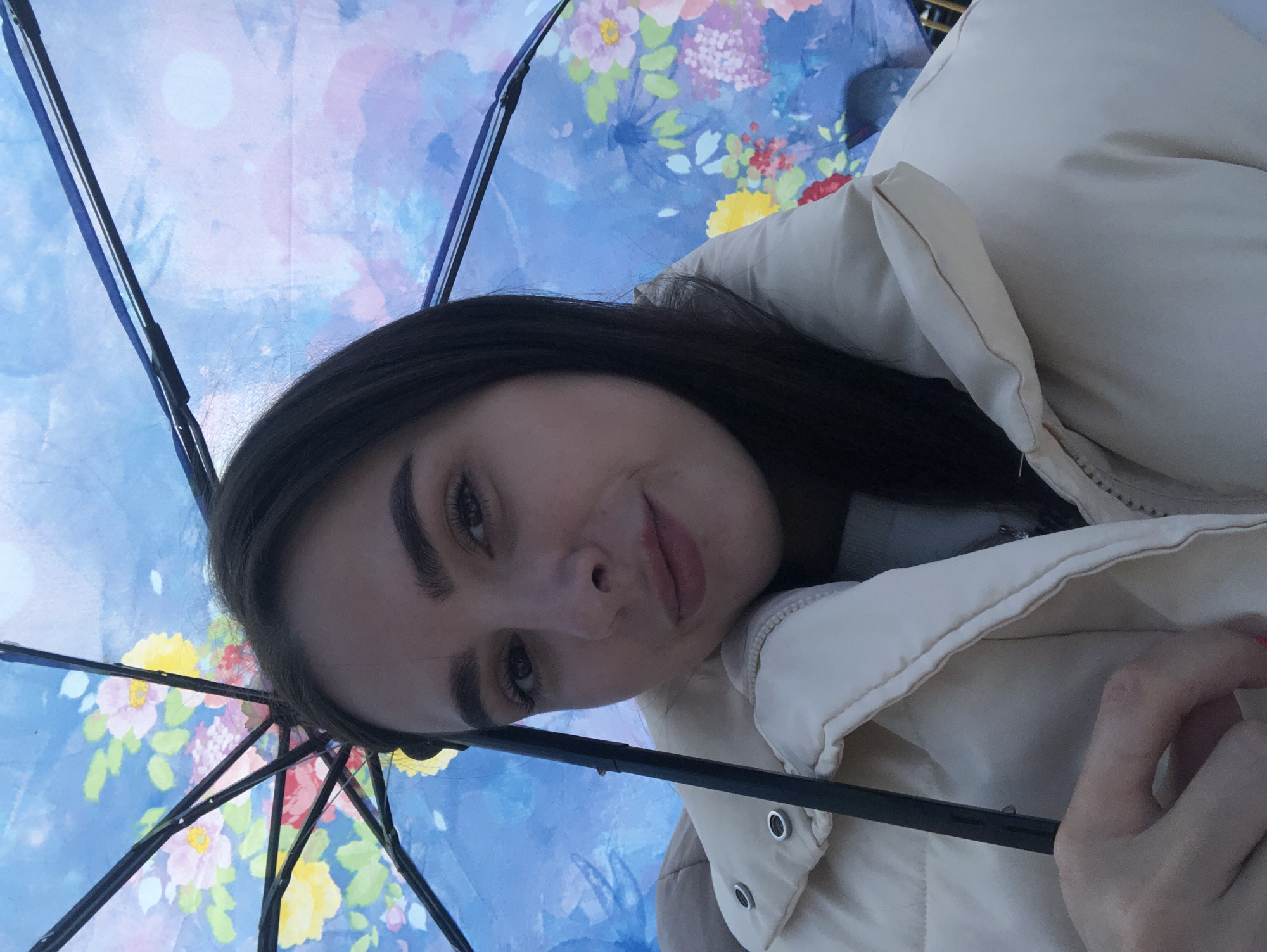 A person with dark hair, wearing a cream-colored puffer jacket, is positioned under an umbrella with a floral and cloudy sky design. The umbrella's black frame is visible. The setting is identified as Chaikovsky, Russia. The person is looking towards the camera, exhibiting a slight smile. The overall composition suggests a selfie taken outdoors.
